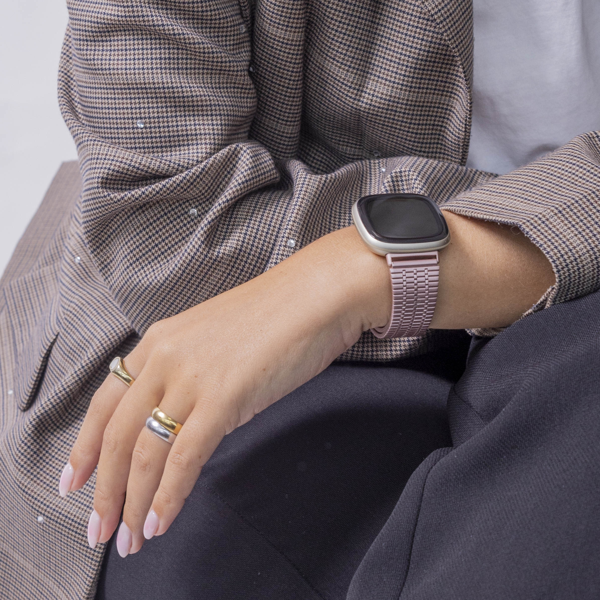Hand mit Uhr. Person trägt Ringe, Blazer und dunkle Kleidung. Neutraler Hintergrund.