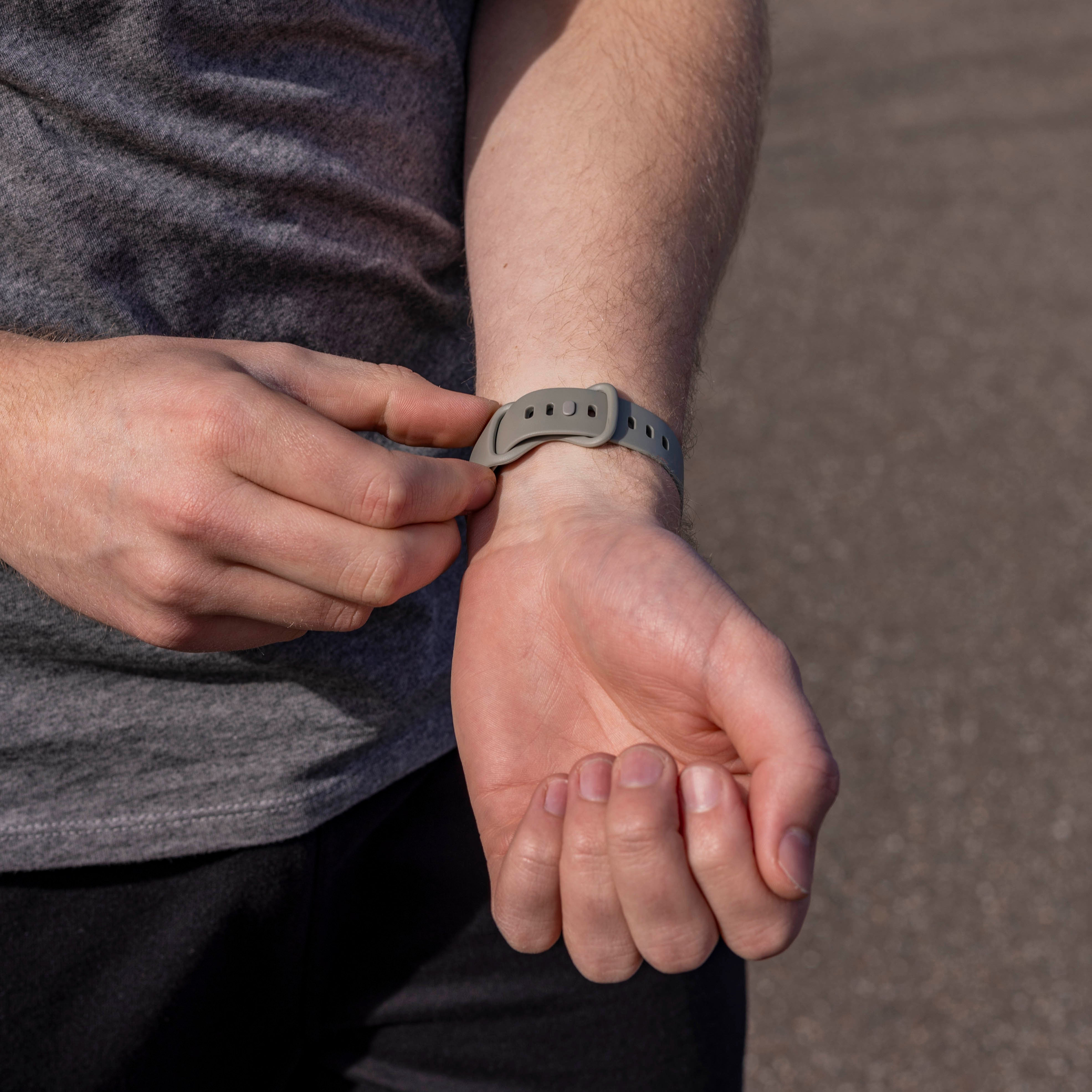 Eine Person, die eine graue Smartwatch an ihrem Handgelenk einstellt. Sie trägt ein graues Hemd, schwarze Hose, und steht auf Asphalt.