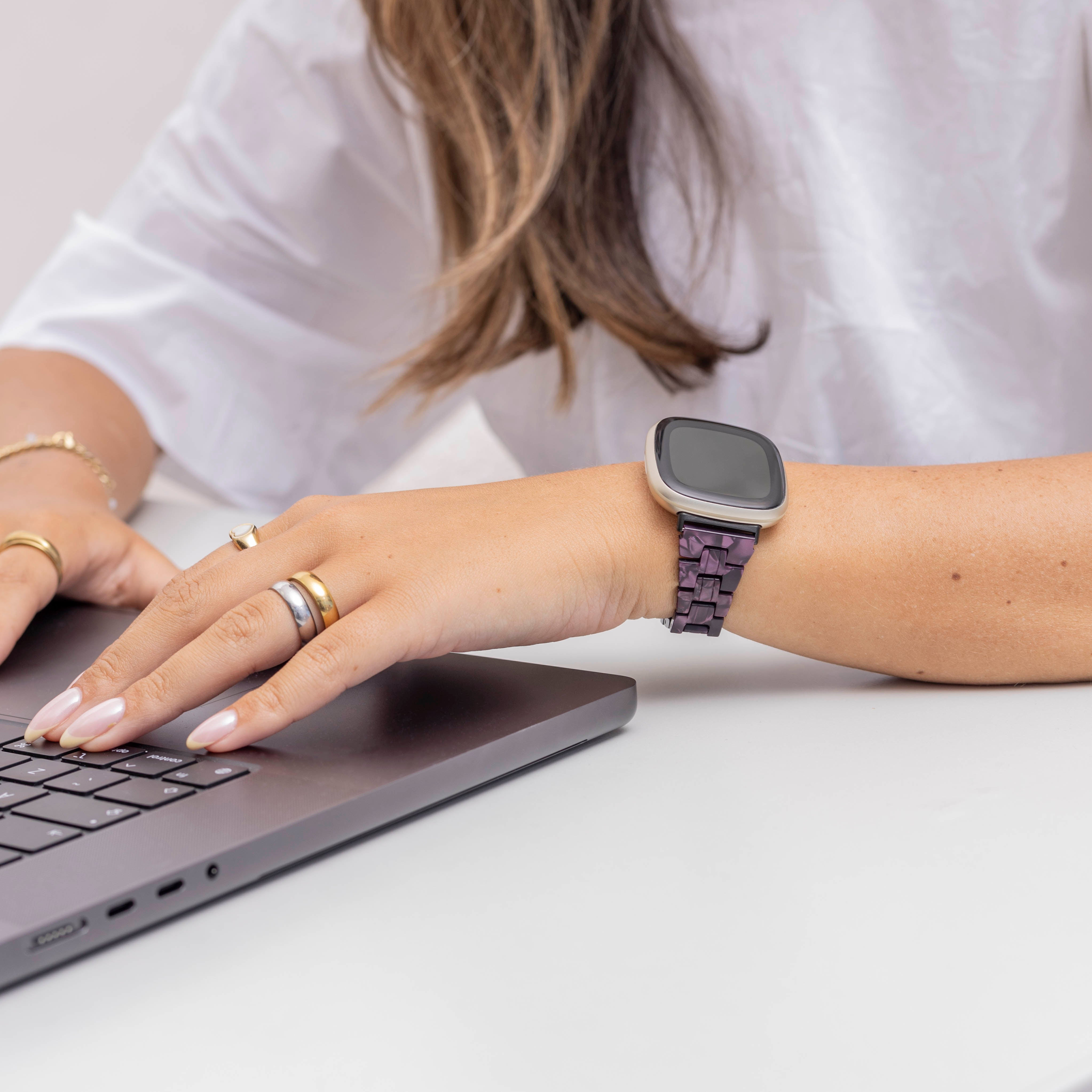 Die Hand einer Frau mit Ringen liegt auf einer Laptop-Tastatur. Eine Smartwatch mit lila Gliedern und ein weißes Hemd sind sichtbar.