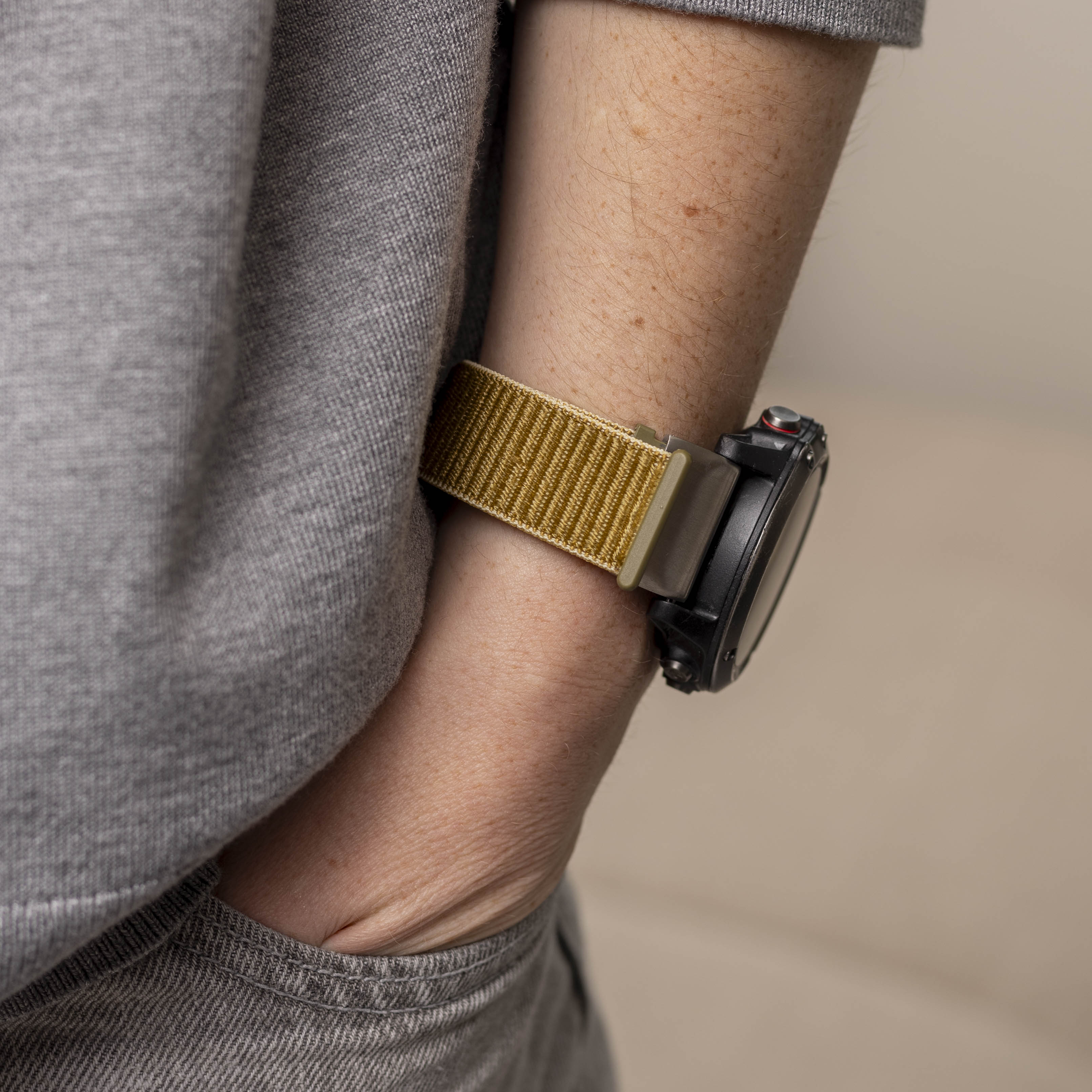 Nahaufnahme eines Arms mit einer Uhr mit goldenem Armband. Der Arm ist vor einem grauen Hemd und hellbeigem Hintergrund.