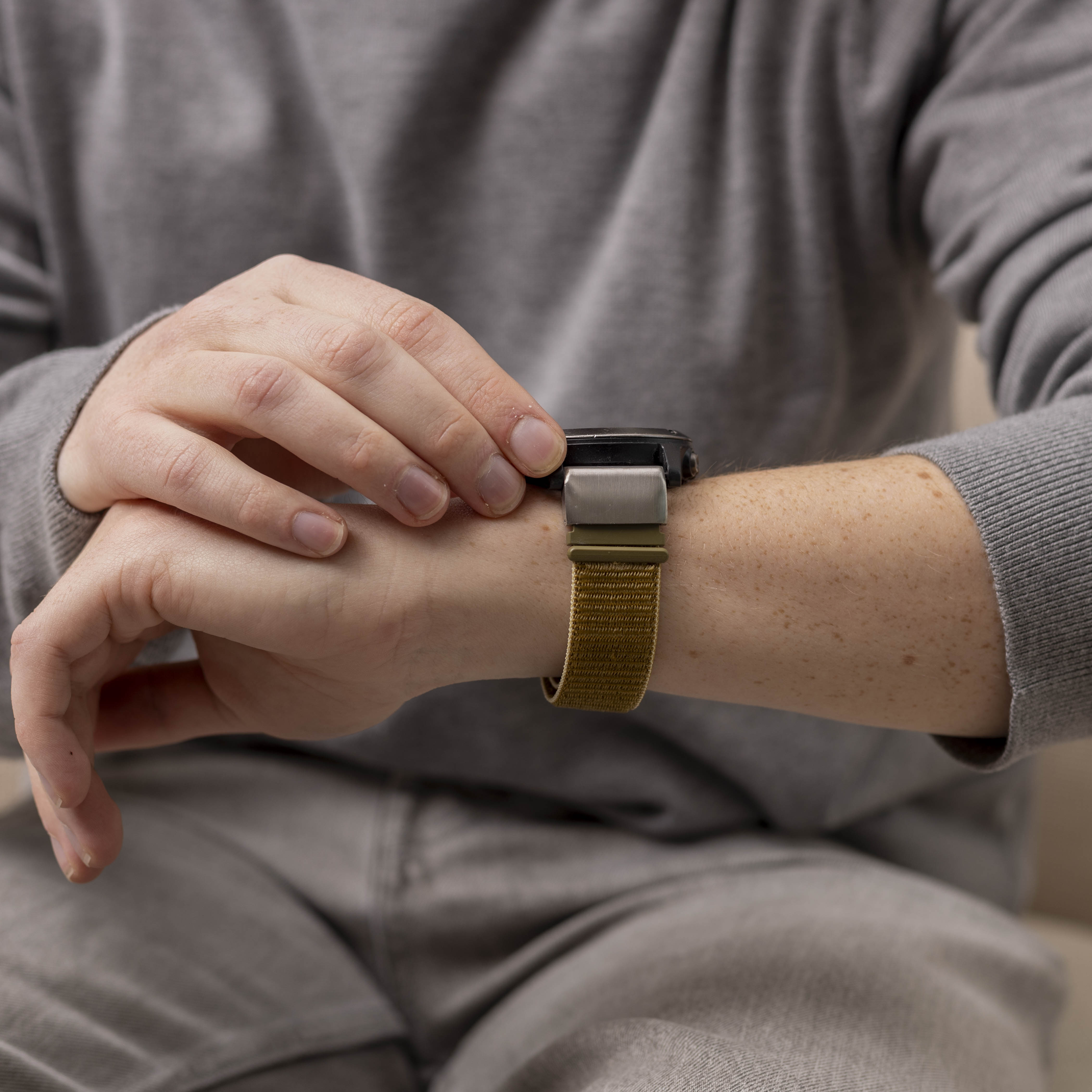 Eine Person in grauem Pullover und Jeans überprüft eine Smartwatch mit khakifarbenem Armband.
