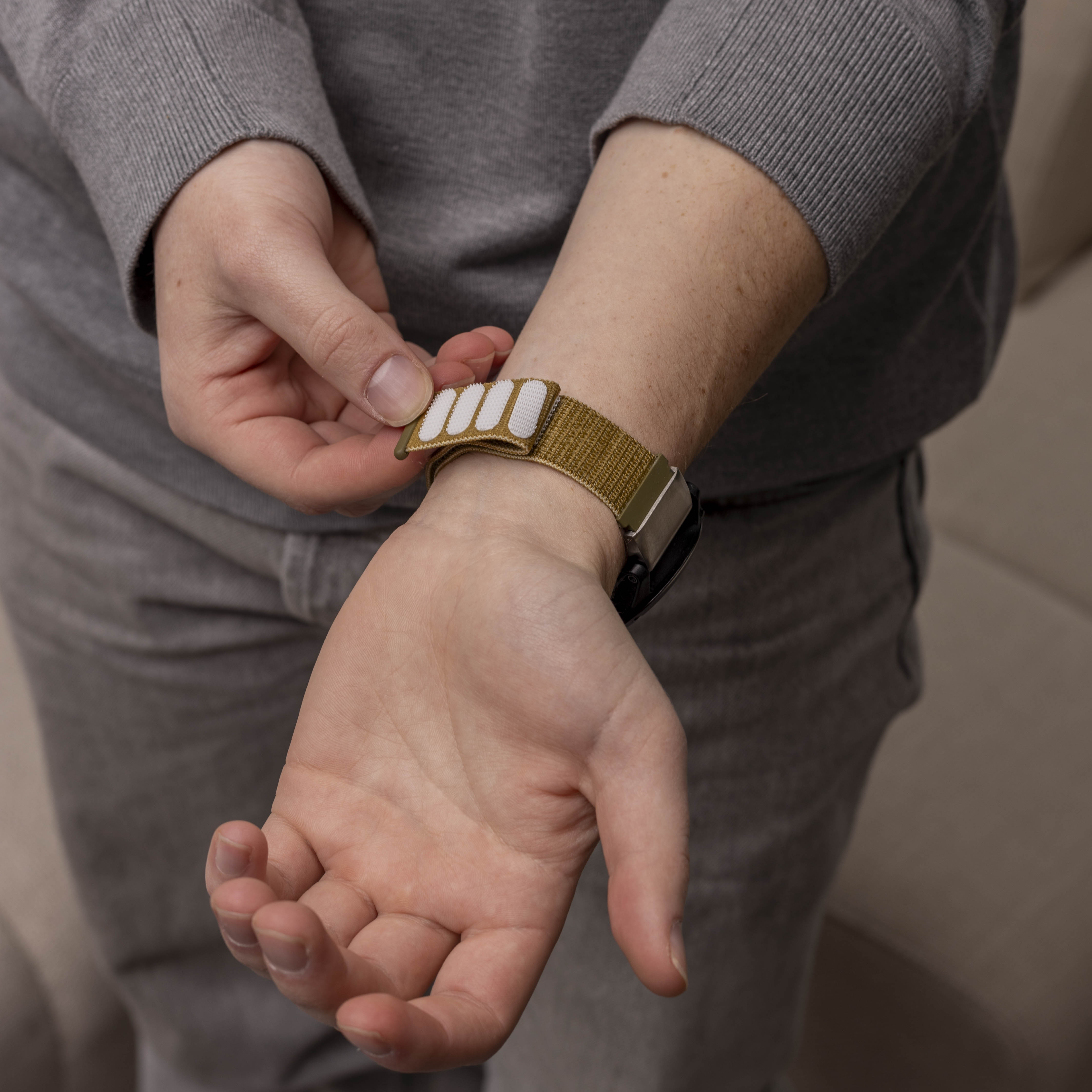 Person passt ein Armband mit weißem Klettverschluss an. Das Band ist olivgrün. Die Person trägt einen grauen Pullover und Hose.