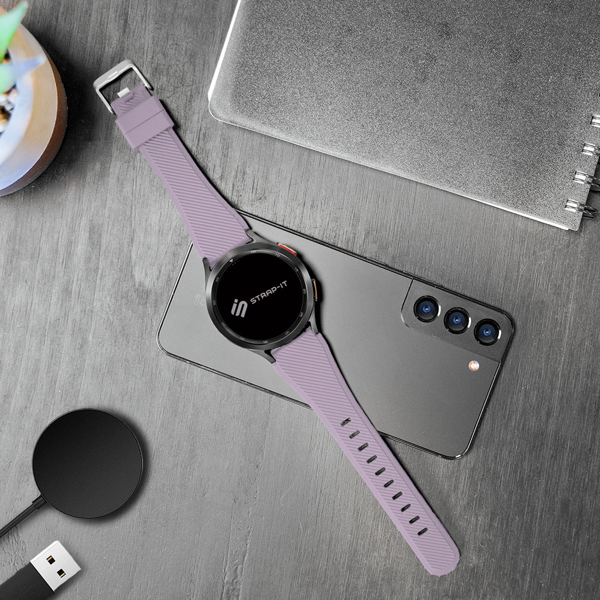 Eine lavendelfarbene Smartwatch mit passendem Armband liegt auf einem grauen Telefon. Ein kabelloses Ladegerät und ein Notizbuch sind ebenfalls vorhanden.
