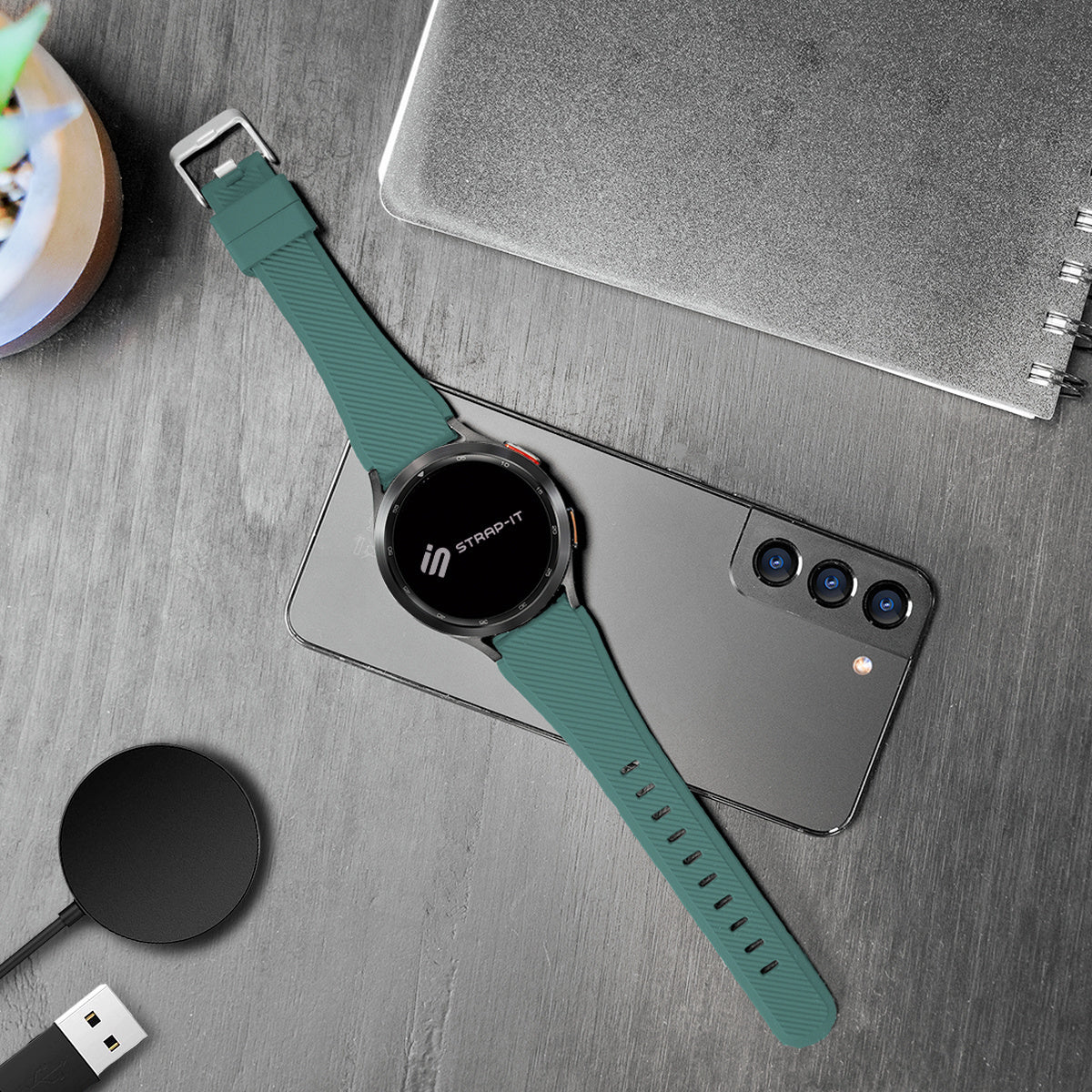 Eine Smartwatch mit grünem Armband liegt auf einem Telefon. Daneben befinden sich ein kabelloses Ladegerät, ein Notizbuch und eine kleine Pflanze.