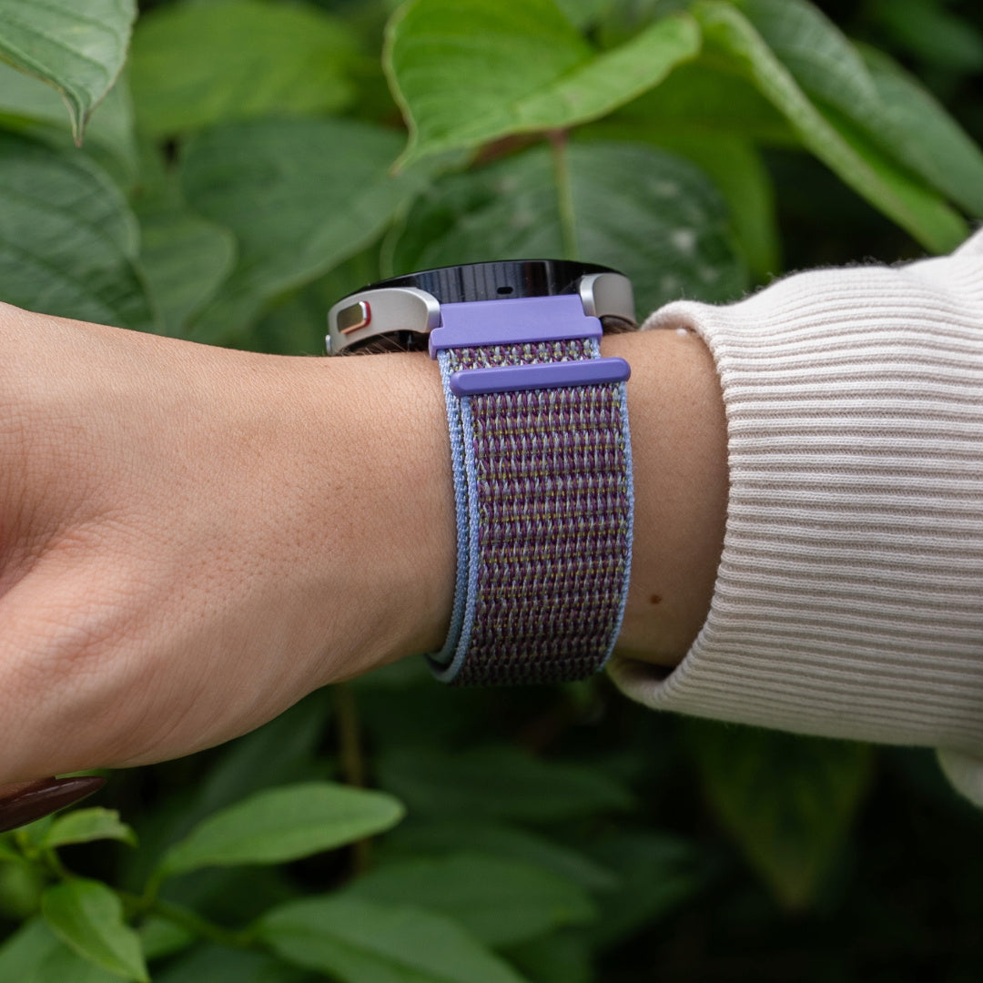 Eine Armbanduhr mit lila Armband an einem Handgelenk. Im Hintergrund grüne Blätter und ein weißes Hemd.