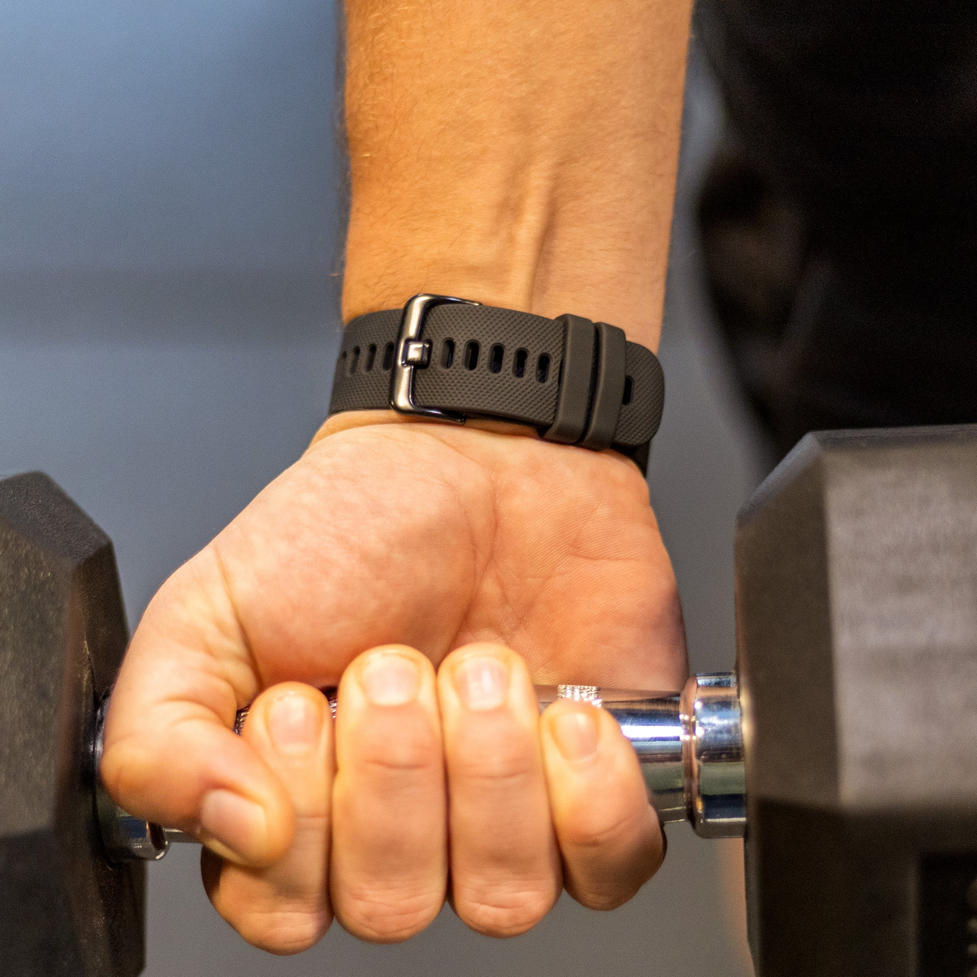 Eine Hand hält eine Hantel. Am Handgelenk befindet sich eine schwarze Uhr. Der Hintergrund ist ein Fitnessstudio.