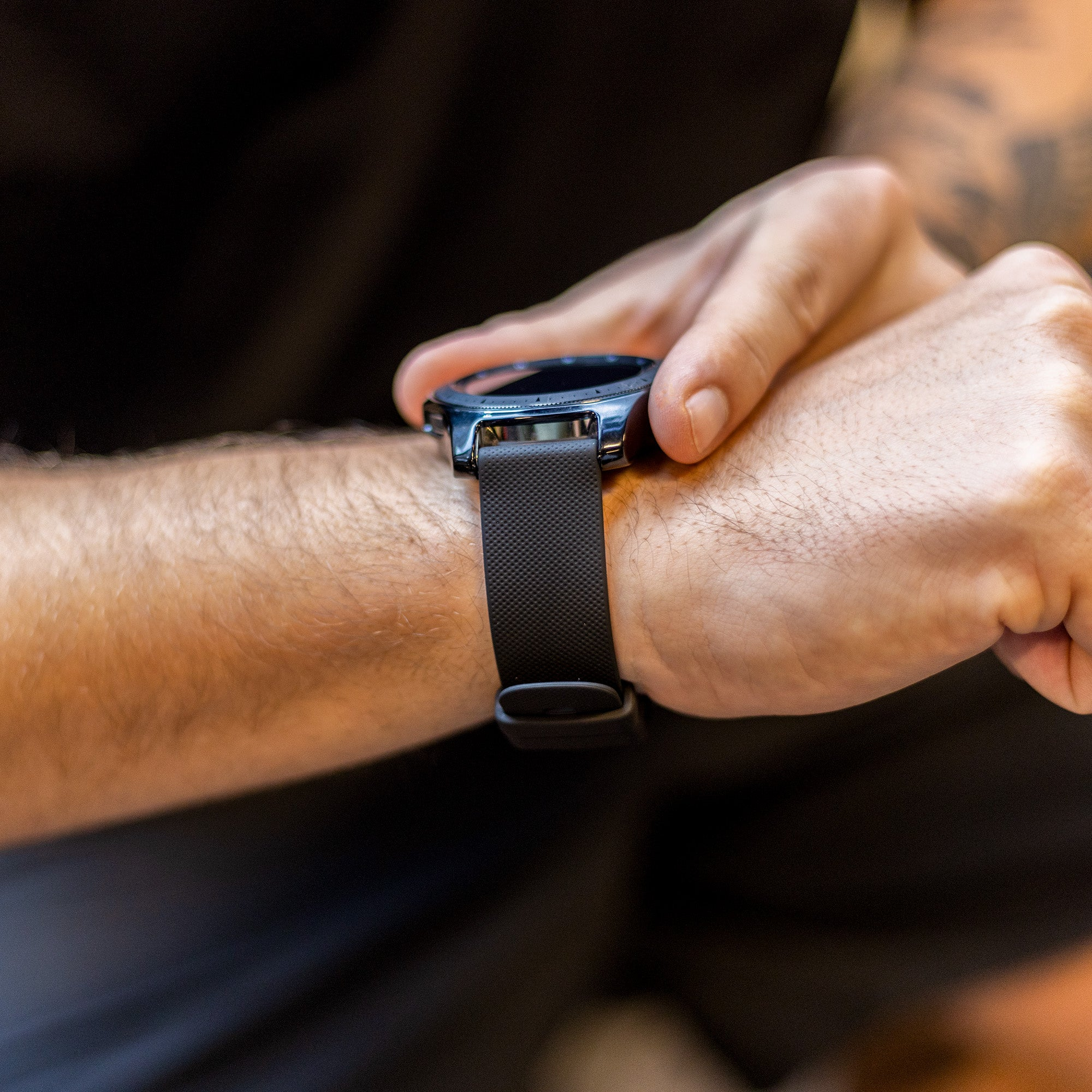 Eine Person, die eine schwarze Uhr mit dunklem Armband trägt und die Zeit abliest. Der Hintergrund ist verschwommen, dunkle Kleidung sichtbar.