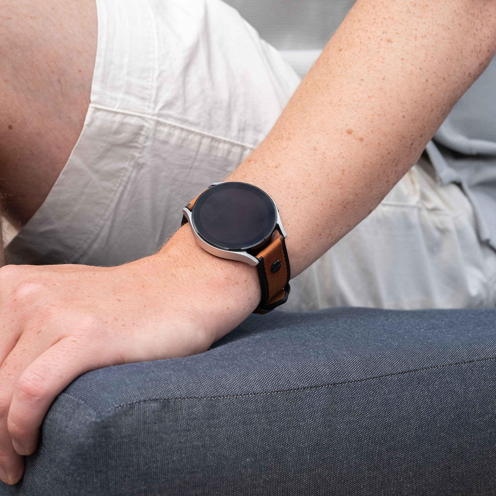Der Arm einer Person mit einer Smartwatch. Die Uhr hat einen schwarzen Bildschirm und ein braunes Lederarmband. Die Person trägt ein weißes Hemd.