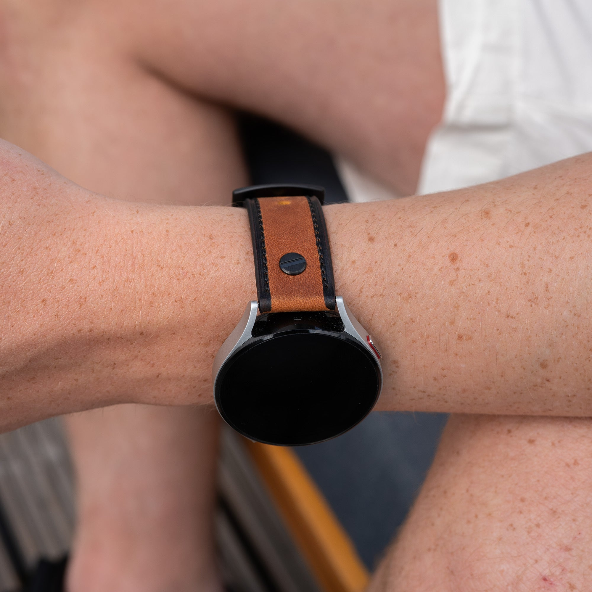 Ein Arm mit einer Uhr. Die Uhr hat einen schwarzen Bildschirm und ein braunes Lederarmband. Die Haut hat Sommersprossen.