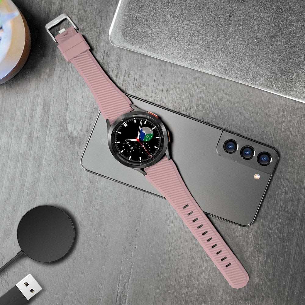 Eine Smartwatch mit pinkfarbenem Armband liegt auf einem Telefon. Ein Ladegerät und ein Laptop sind ebenfalls zu sehen.
