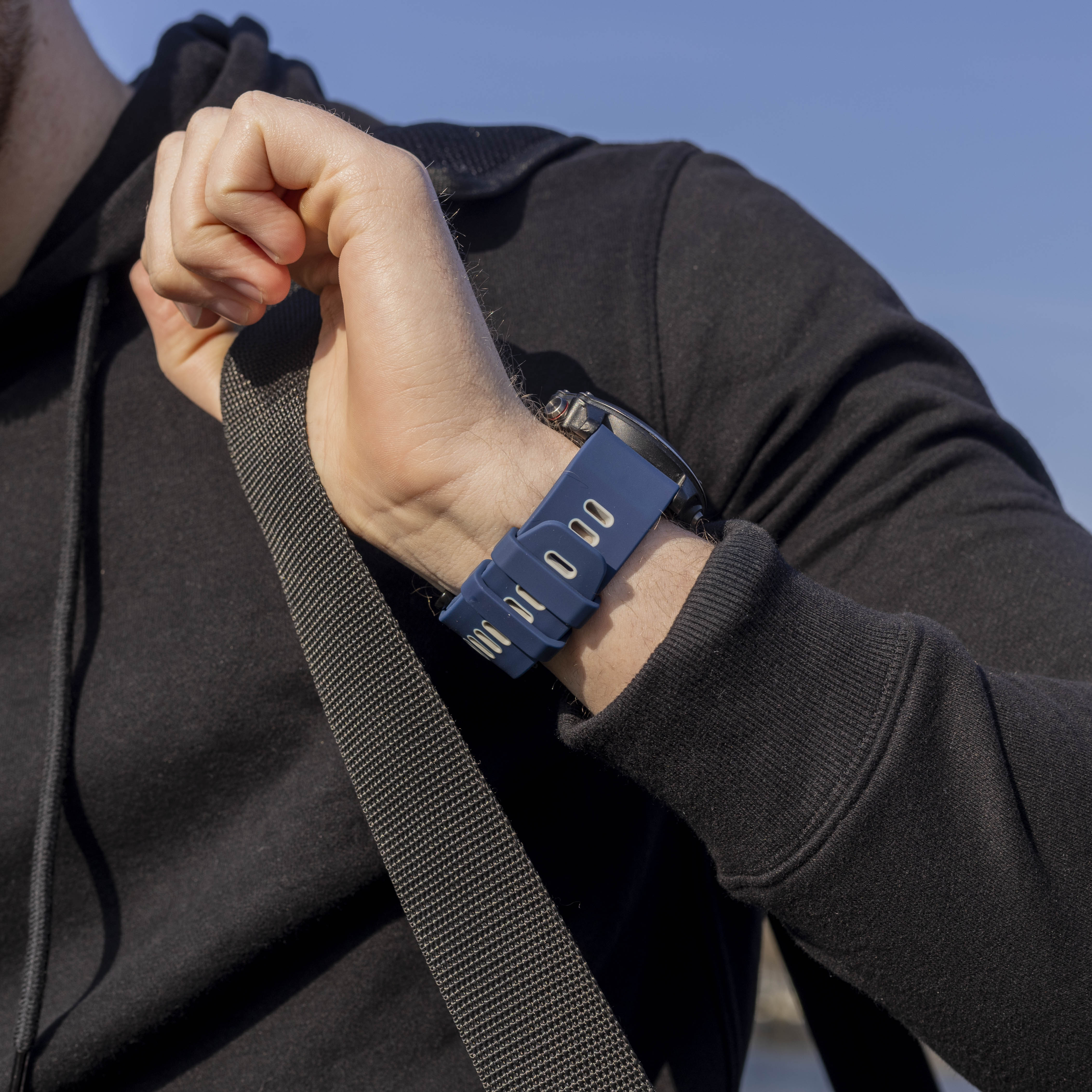 Eine Person trägt einen schwarzen Hoodie und eine Uhr mit blauem Armband. Ein schwarzer Taschenriemen ist sichtbar.