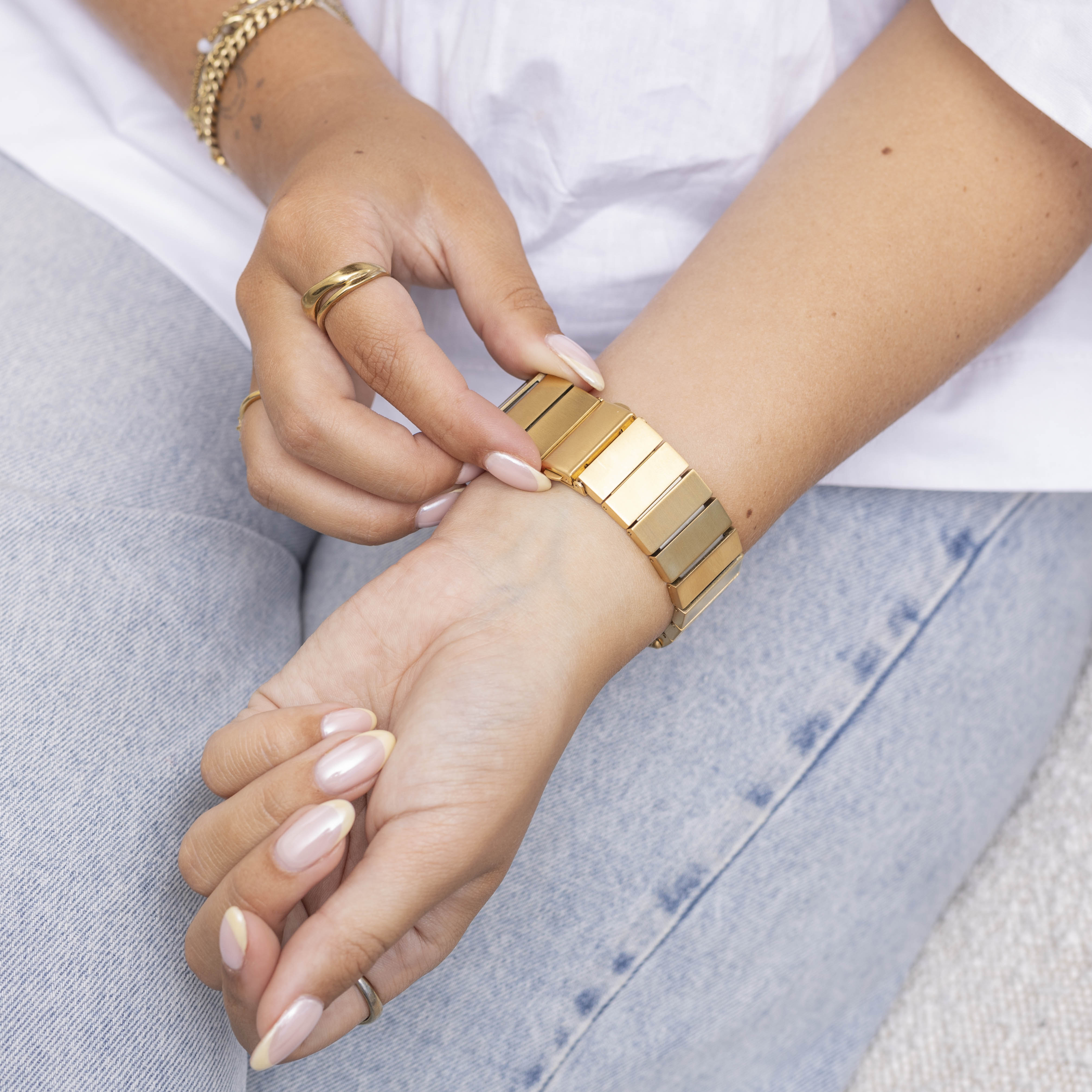 Hände einer Person mit Ringen und einem goldenen Armband am Handgelenk. Sie trägt auch ein weißes Hemd und Jeans.
