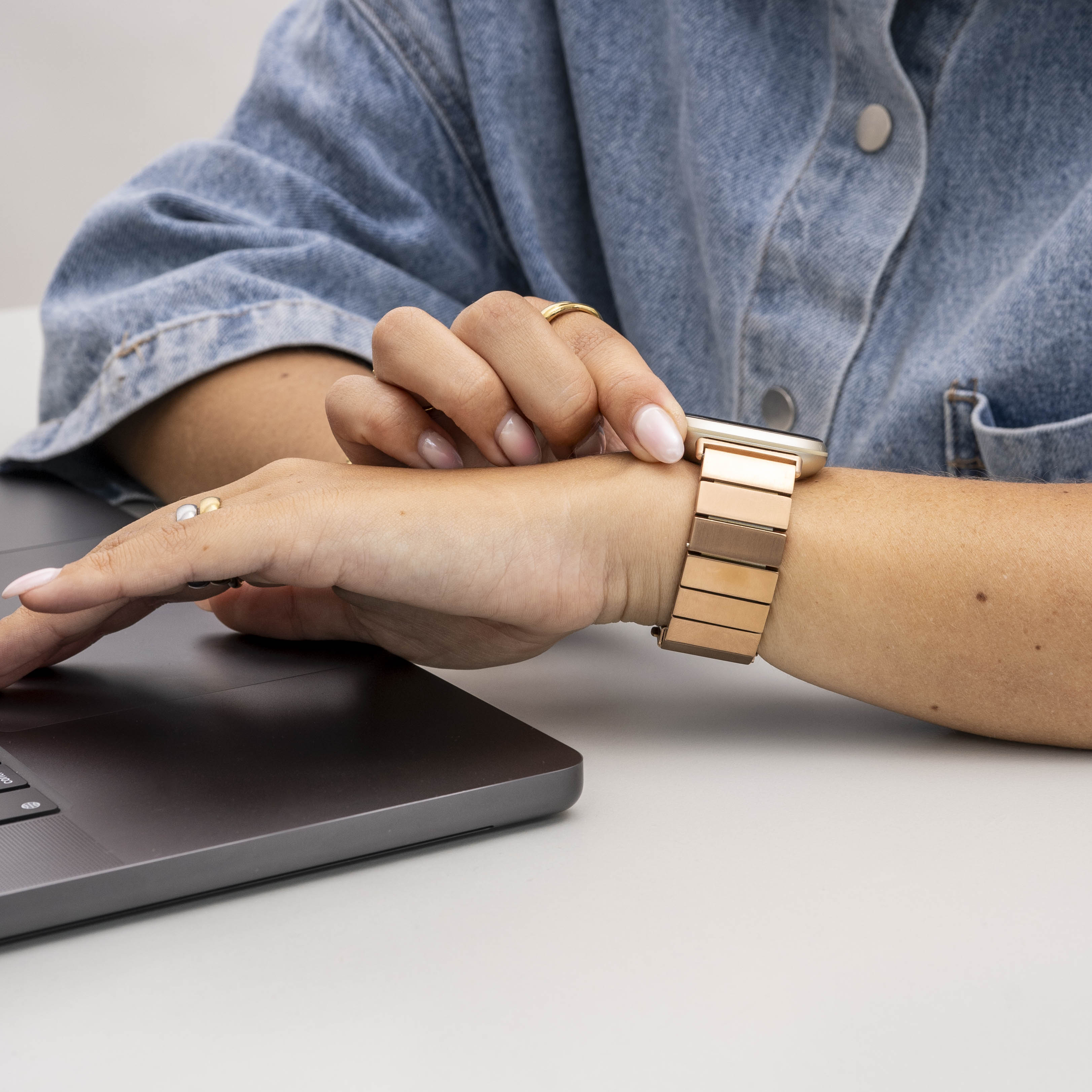 Eine Person in einem Jeanshemd benutzt einen Laptop. Sie trägt eine goldene Uhr und Ringe. Der Hintergrund ist weiß.