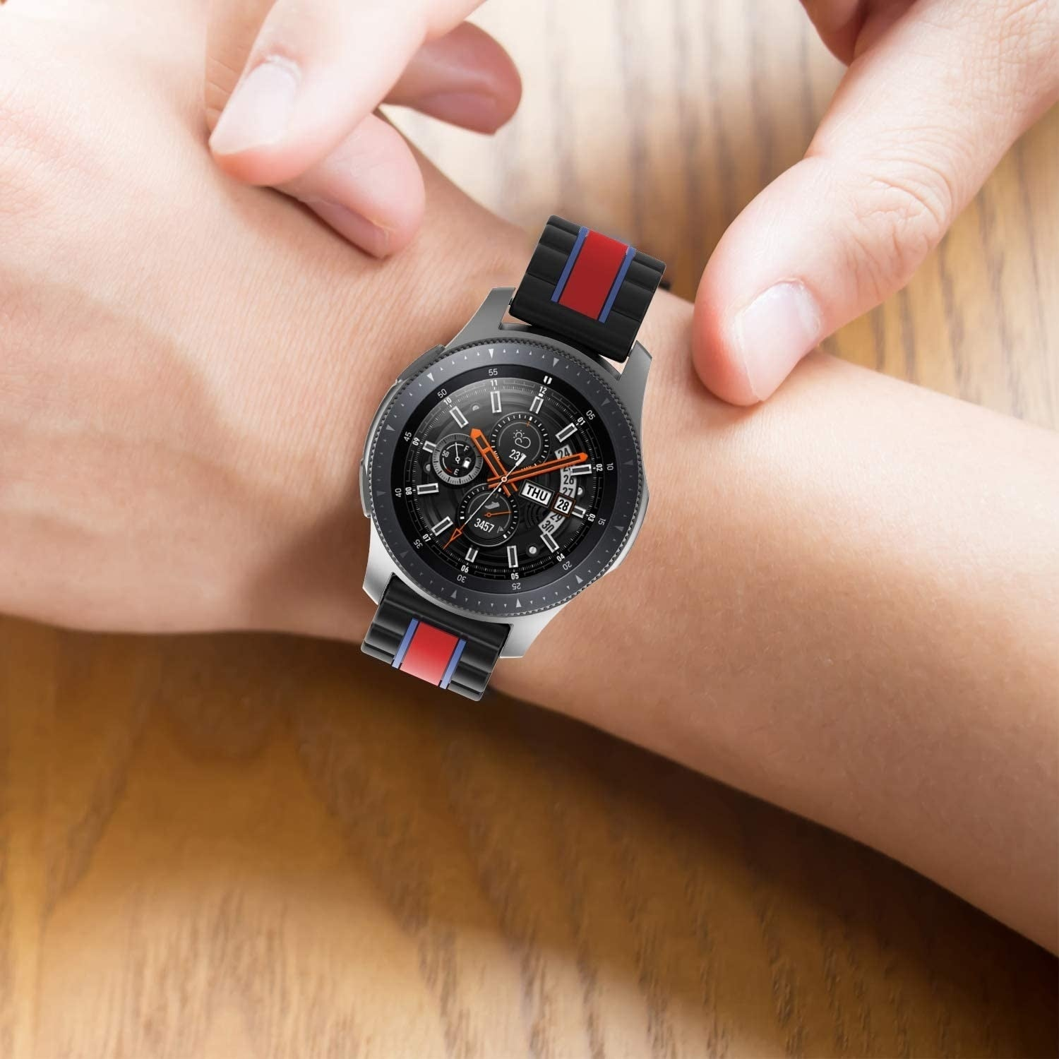 Handgelenk mit Smartwatch. Die Uhr hat ein schwarzes Zifferblatt, eine silberne Lünette und ein schwarzes Armband mit roten und blauen Streifen.