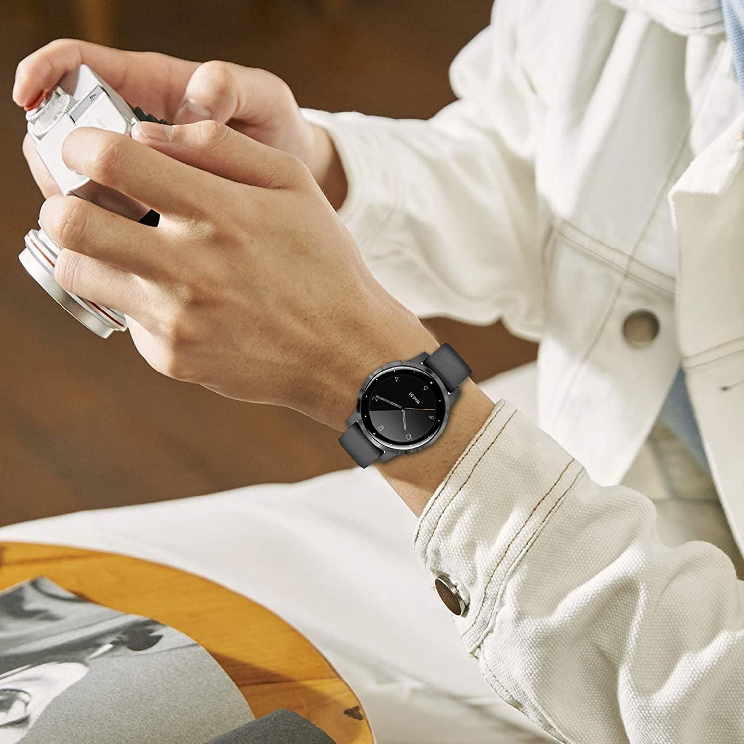 Eine Person mit Smartwatch und Kamera. Sie trägt eine weiße Jacke. Die Uhr hat ein schwarzes Zifferblatt und graues Armband.