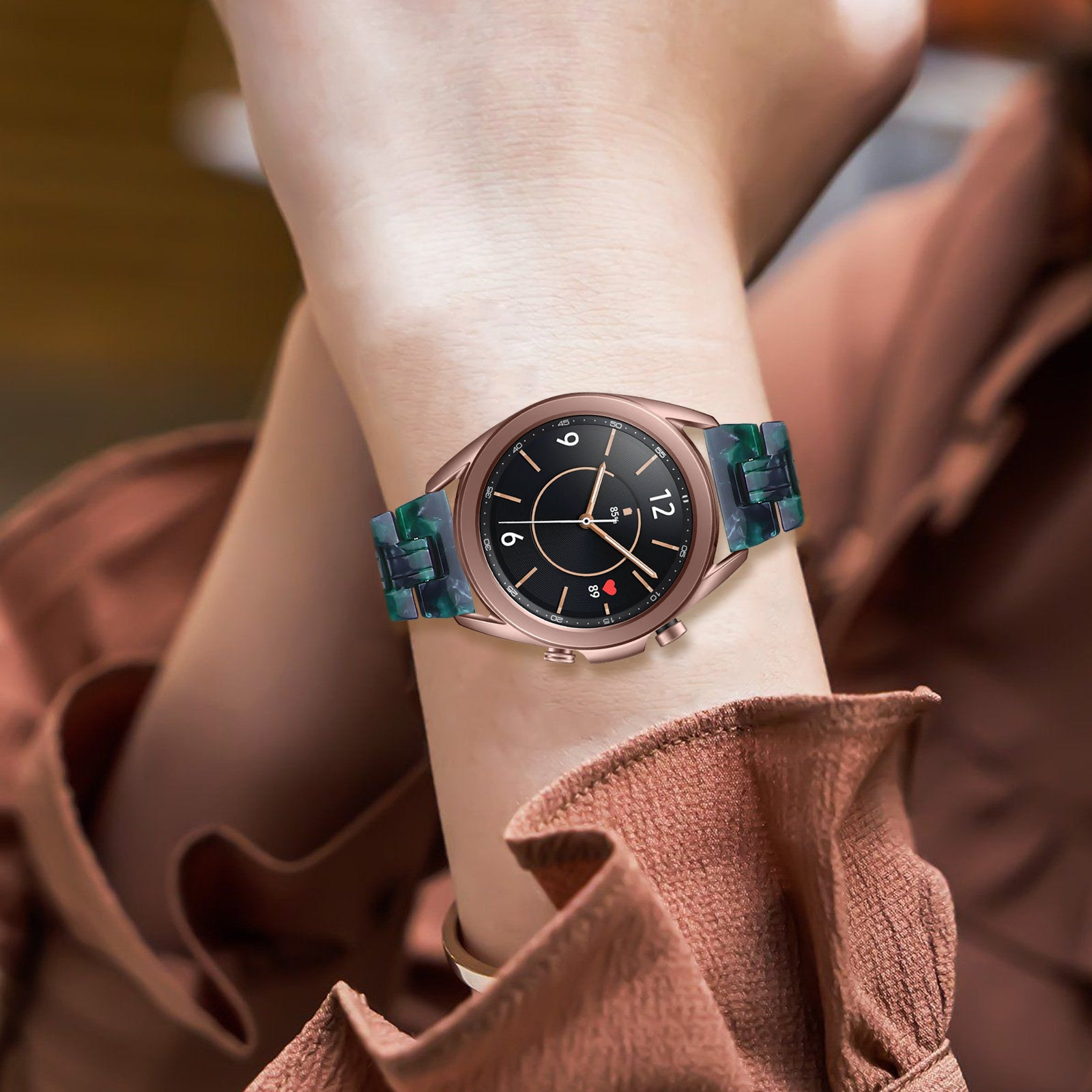 Eine roségoldene Smartwatch mit schwarzem Zifferblatt und grün gemustertem Armband am Handgelenk einer Person. Die Person trägt ein braunes Oberteil.