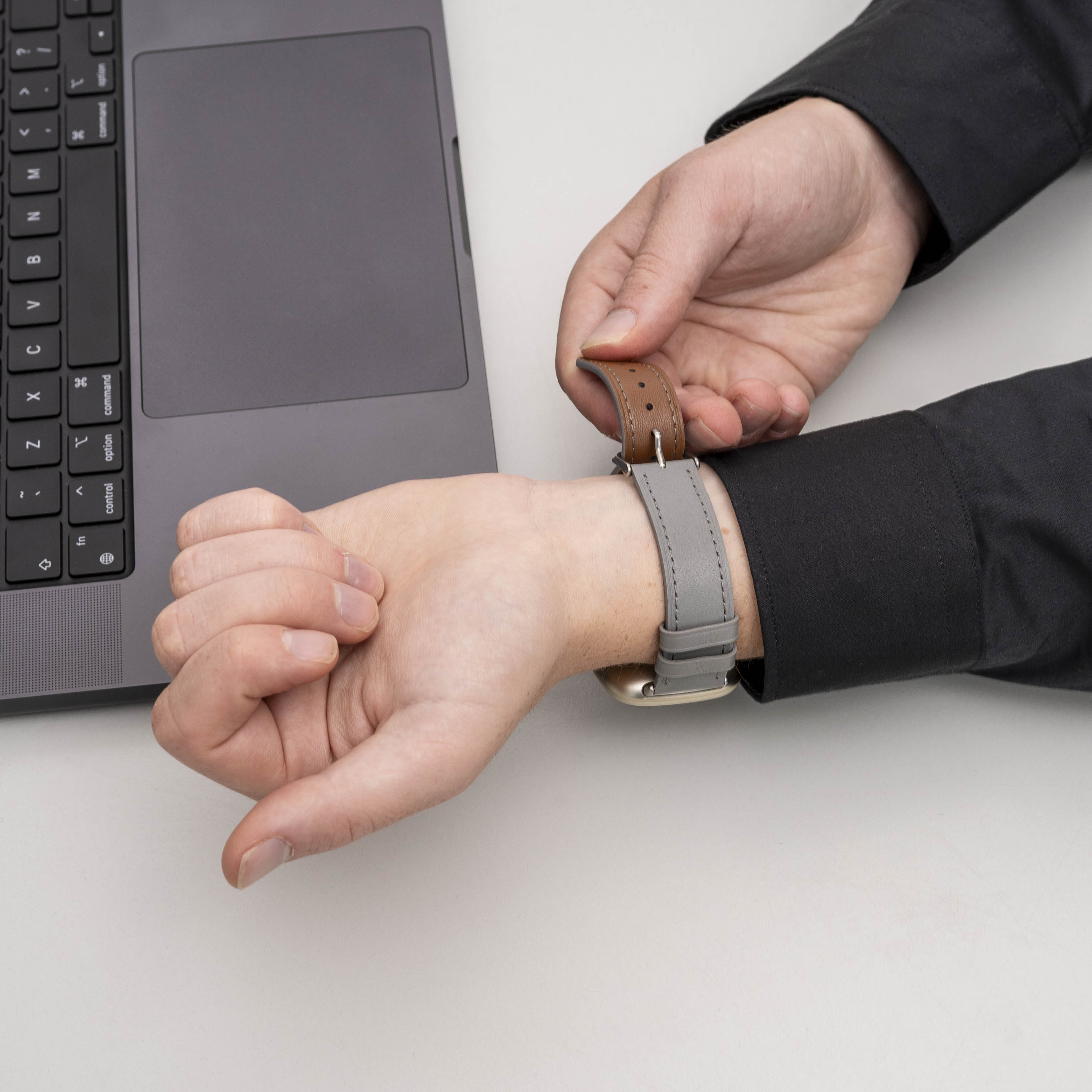 Eine Person befestigt ein Lederarmband an ihrem Handgelenk, neben einem Laptop auf einer weißen Oberfläche.