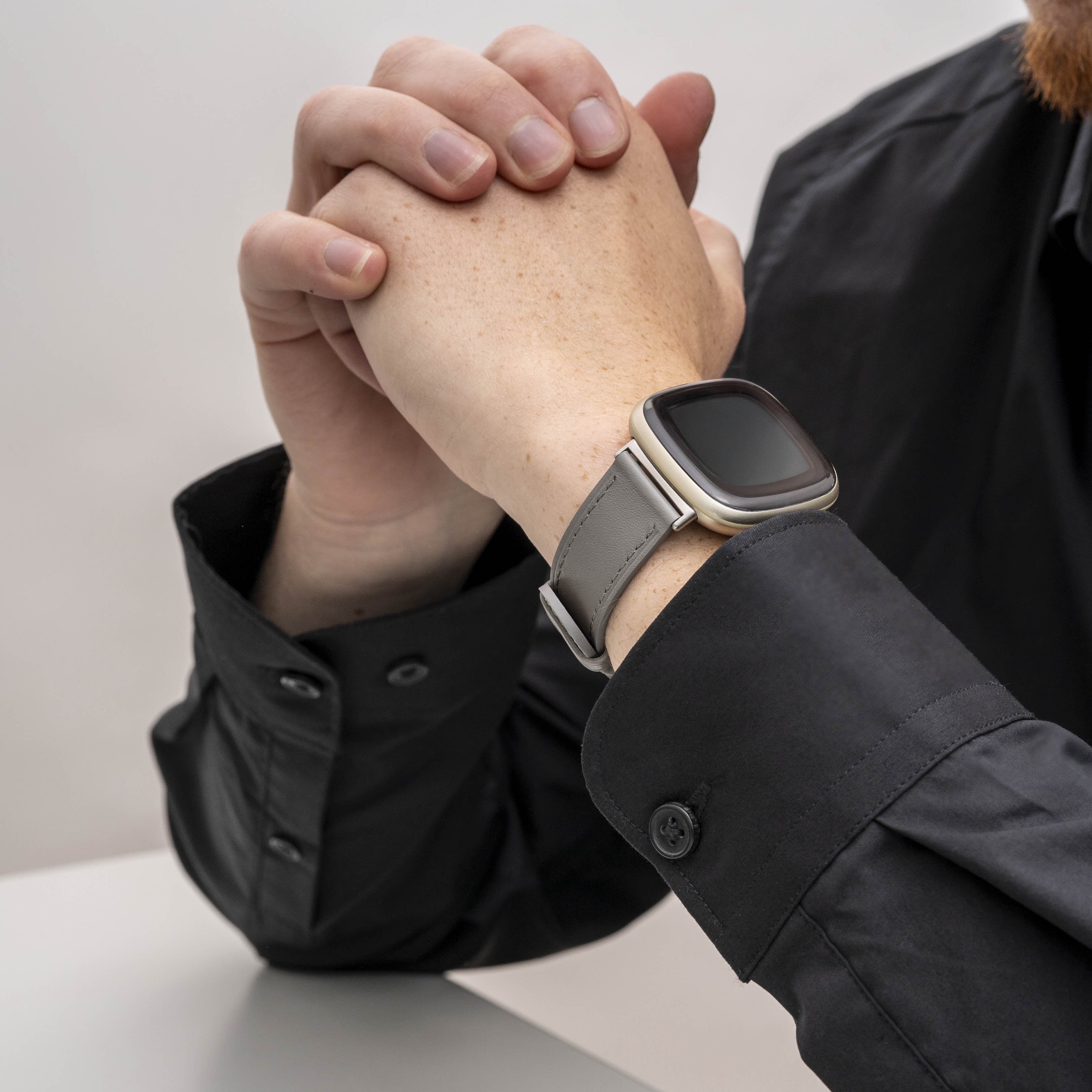 Person trägt ein schwarzes Hemd und eine graue Uhr, Hände gefaltet. Uhr am linken Handgelenk.