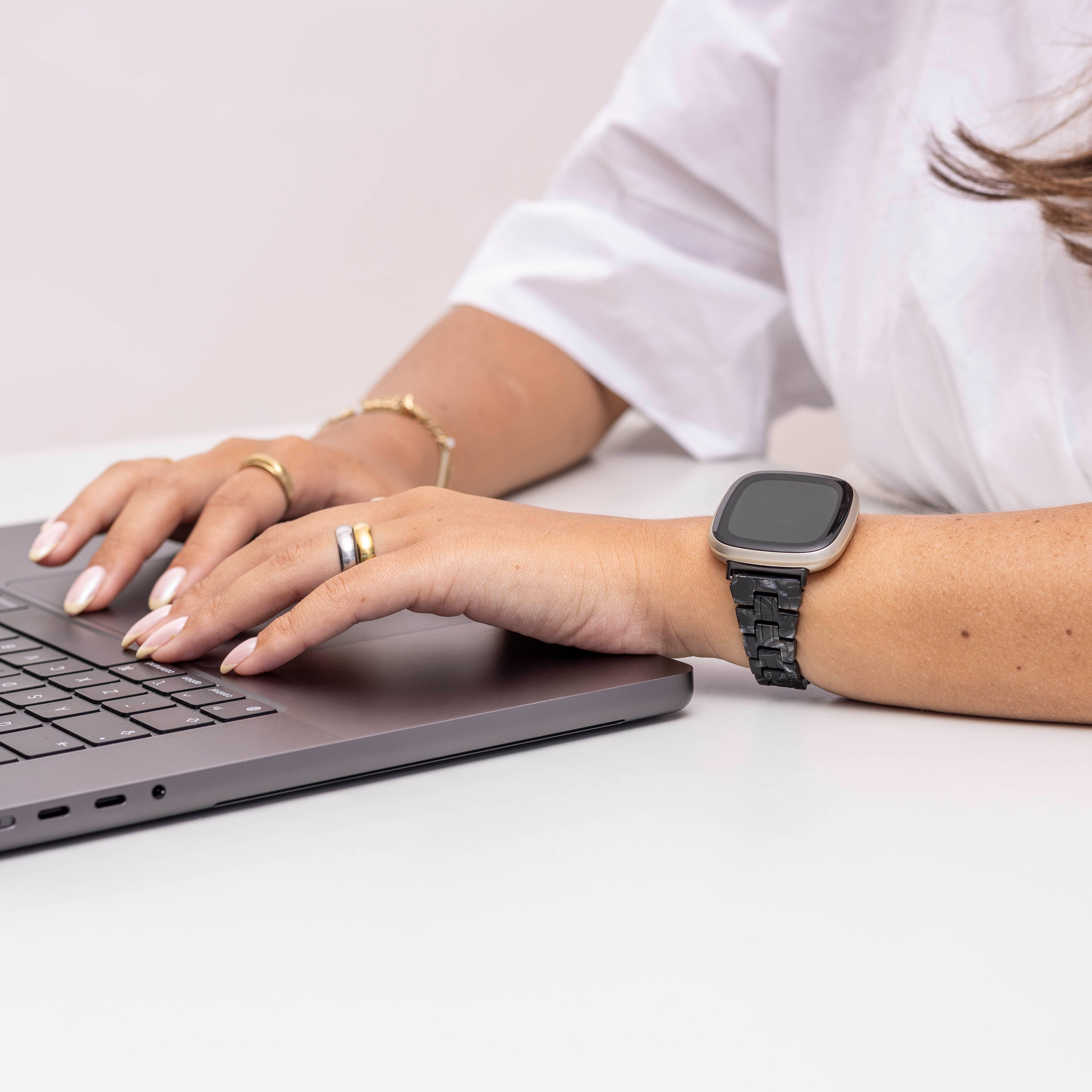 Hände tippen auf einer Laptop-Tastatur. Am Handgelenk ist eine Smartwatch. Sie trägt Ringe und ein weißes Hemd.