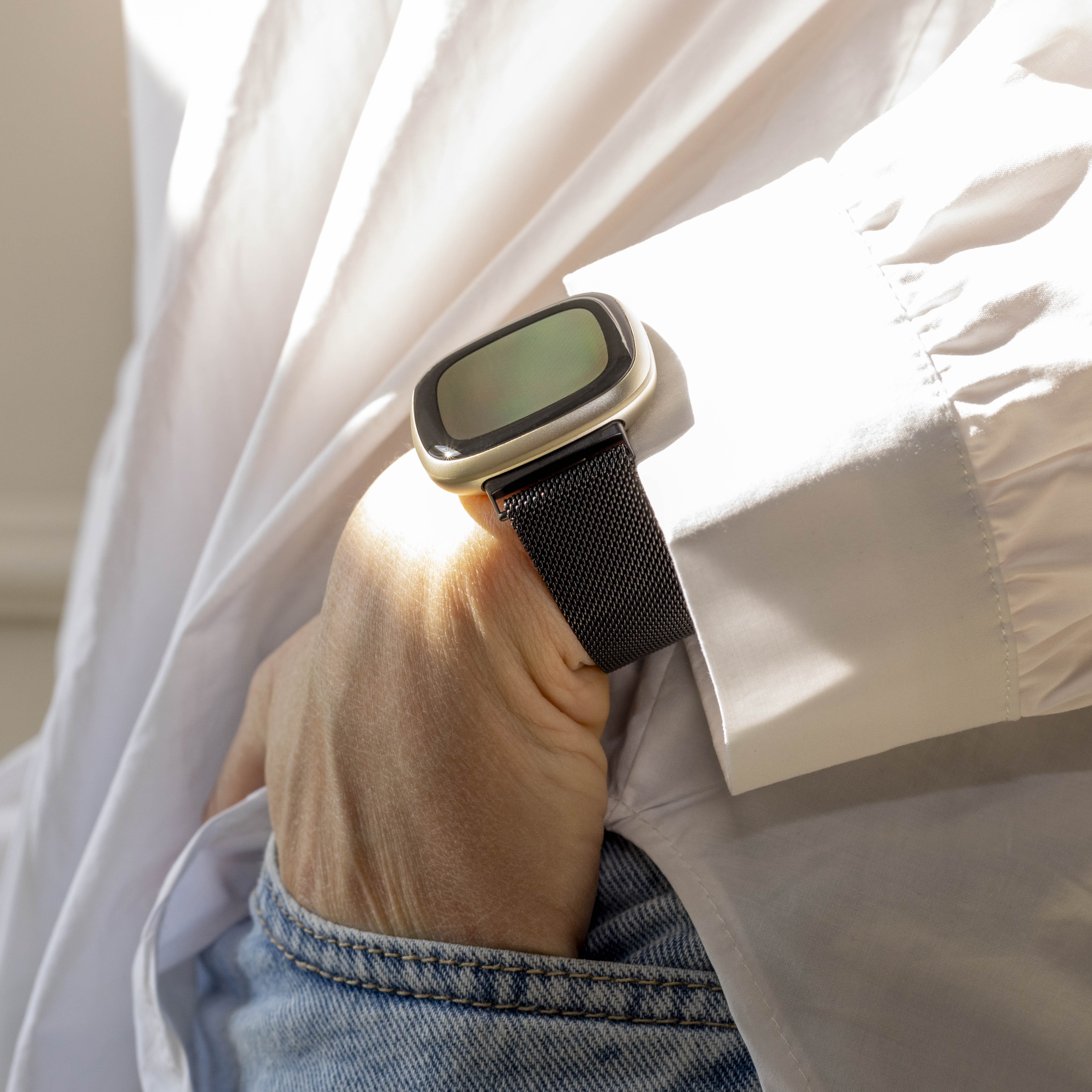 Eine Person mit weißem Hemd und Jeans hält eine Smartwatch. Die Uhr hat ein schwarzes Armband und eine goldene Lünette.