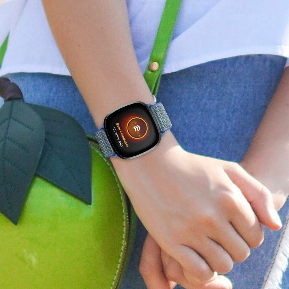 Das Handgelenk einer Person mit einer Smartwatch, die 'Ziel erreicht' anzeigt. Die Uhr hat ein blaues Armband. Im Hintergrund sind eine grüne Tasche und Jeans zu sehen.