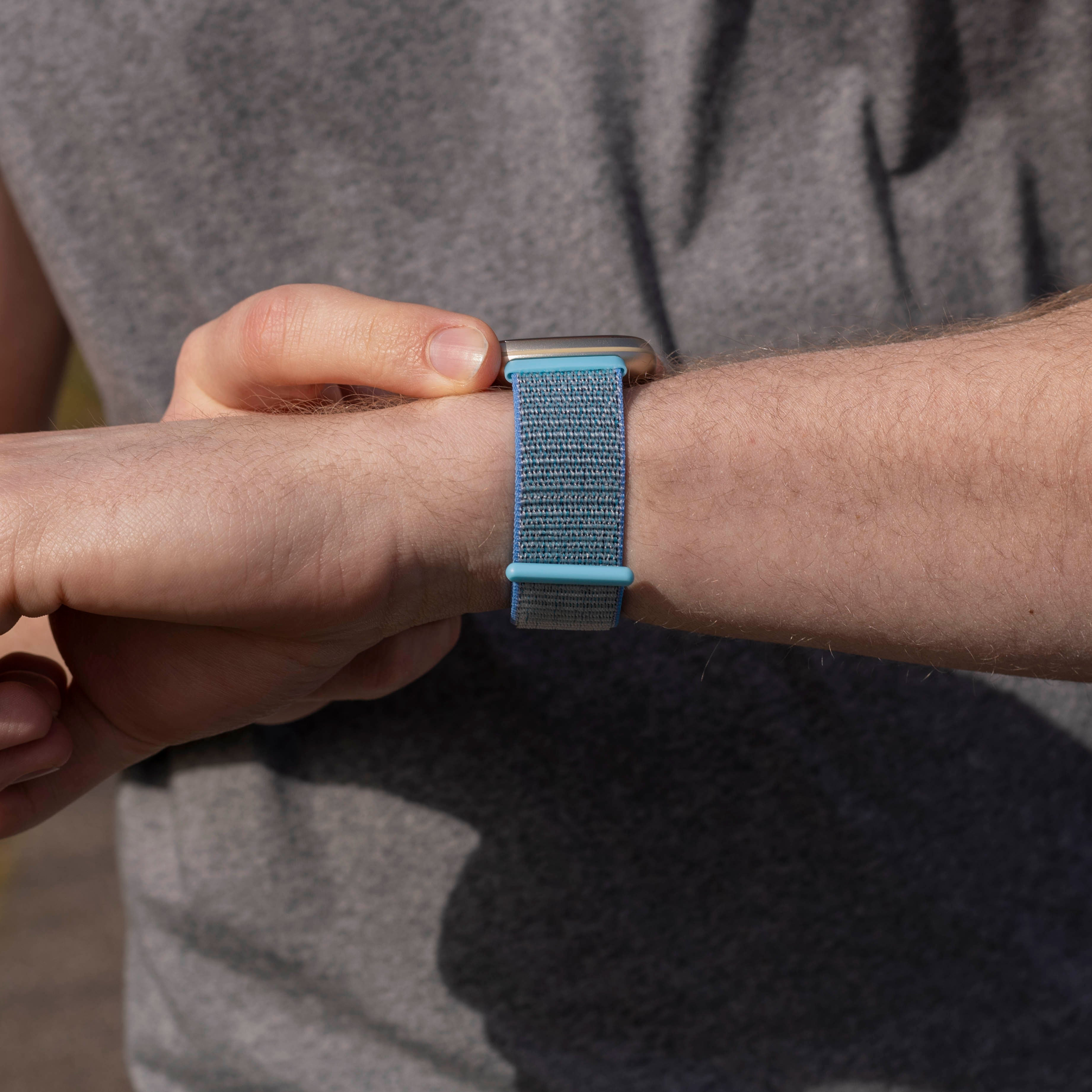 Handgelenk einer Person mit blau-grauem Armband. Person trägt graues Hemd.