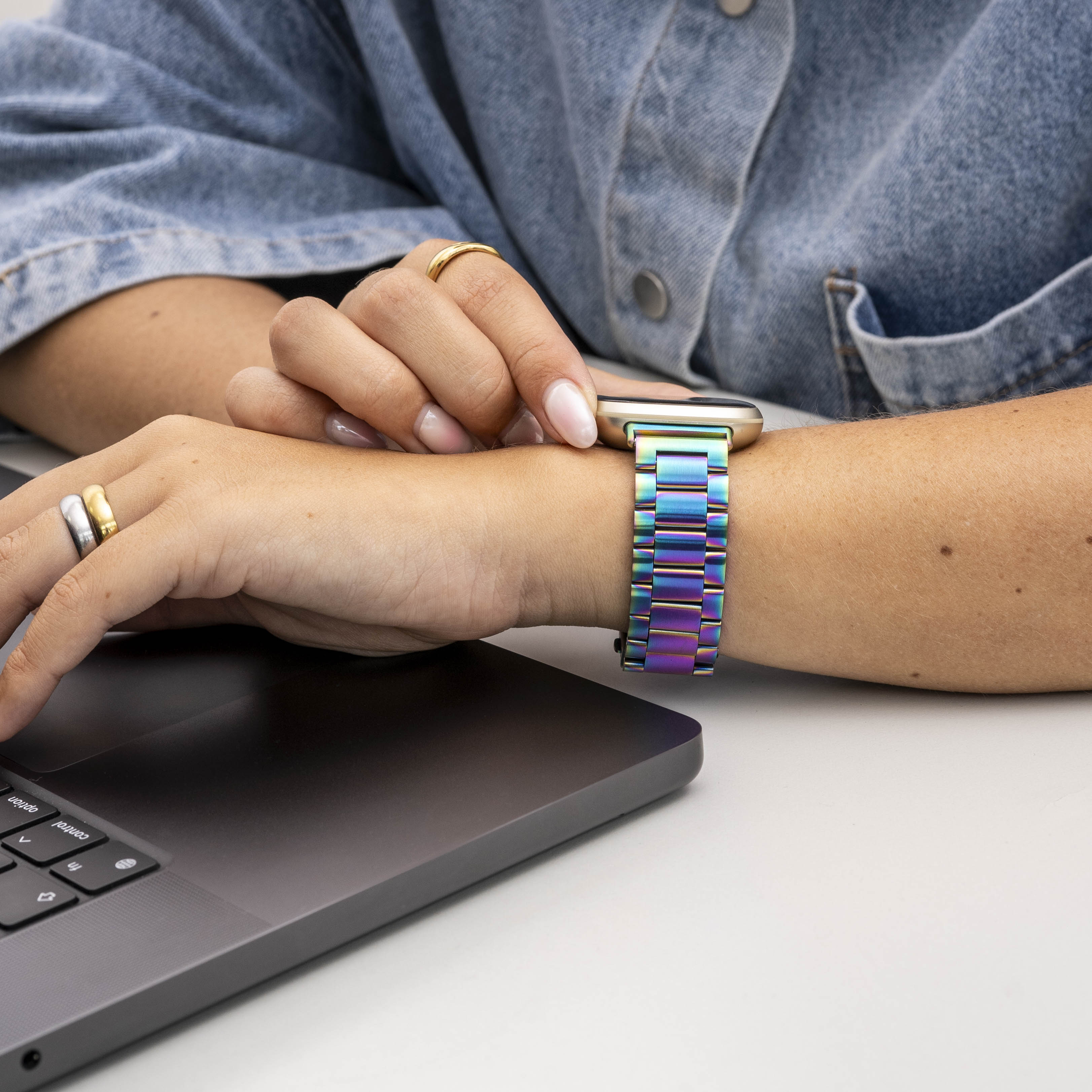 Eine Person trägt ein Jeanshemd und eine Regenbogenuhr. Sie hat Ringe und benutzt einen Laptop auf einer weißen Oberfläche.