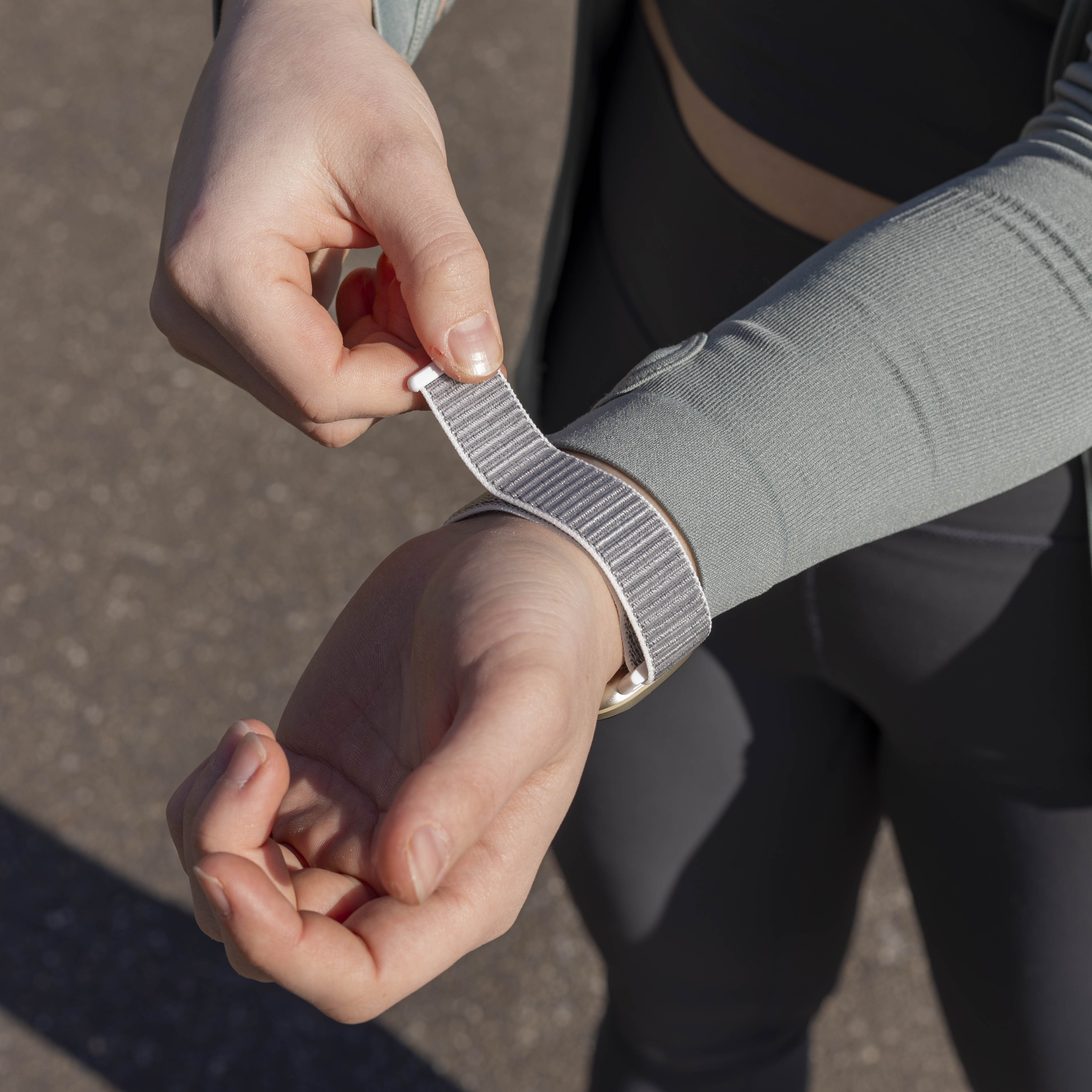 Person passt ein graues Uhrenarmband am Handgelenk an. Sie trägt ein hellgrünes Oberteil. Der Hintergrund ist verschwommener Asphalt.