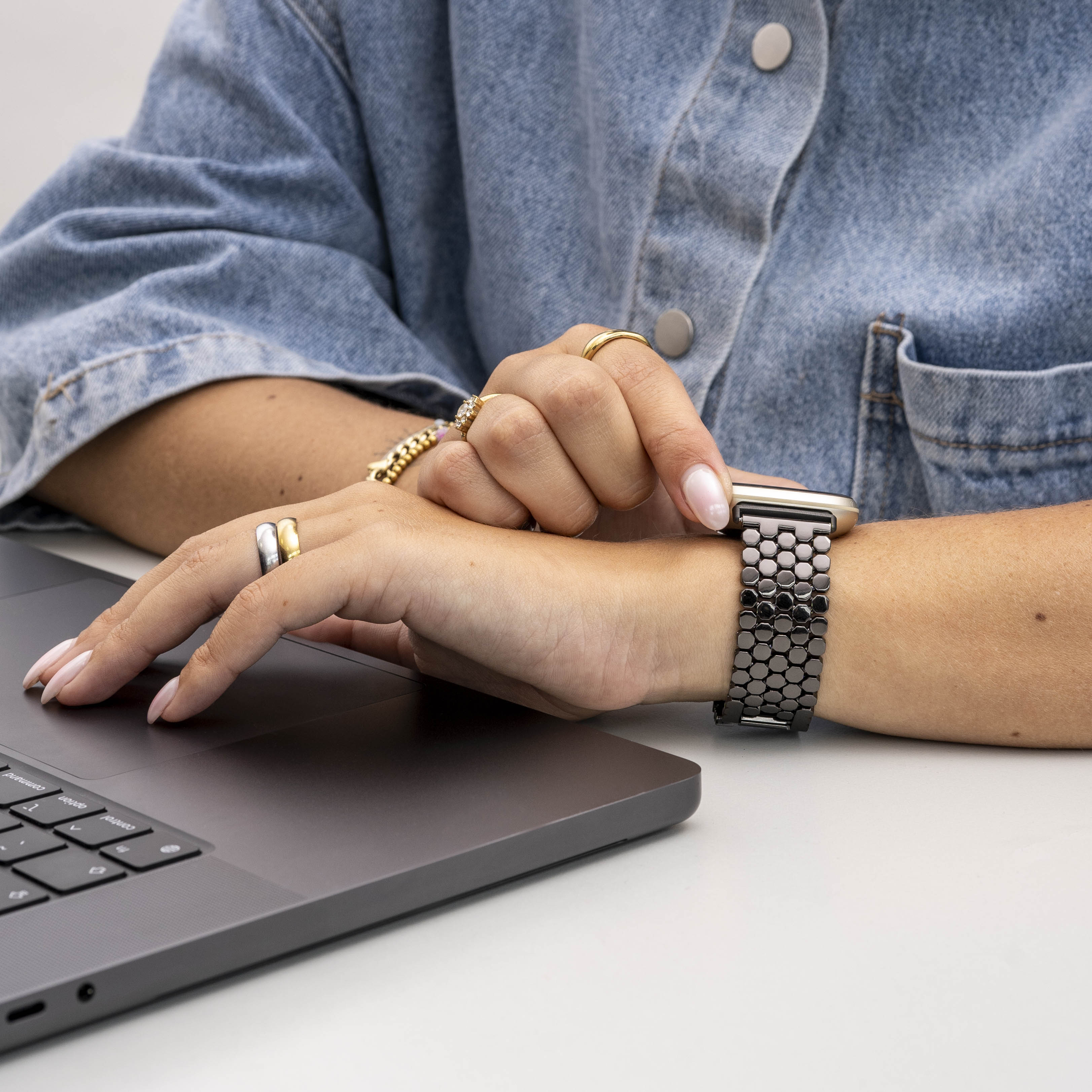 Eine Person mit Jeansjacke, Ringen und einer Uhr mit schwarzem Band, die einen Laptop auf einer weißen Oberfläche benutzt.