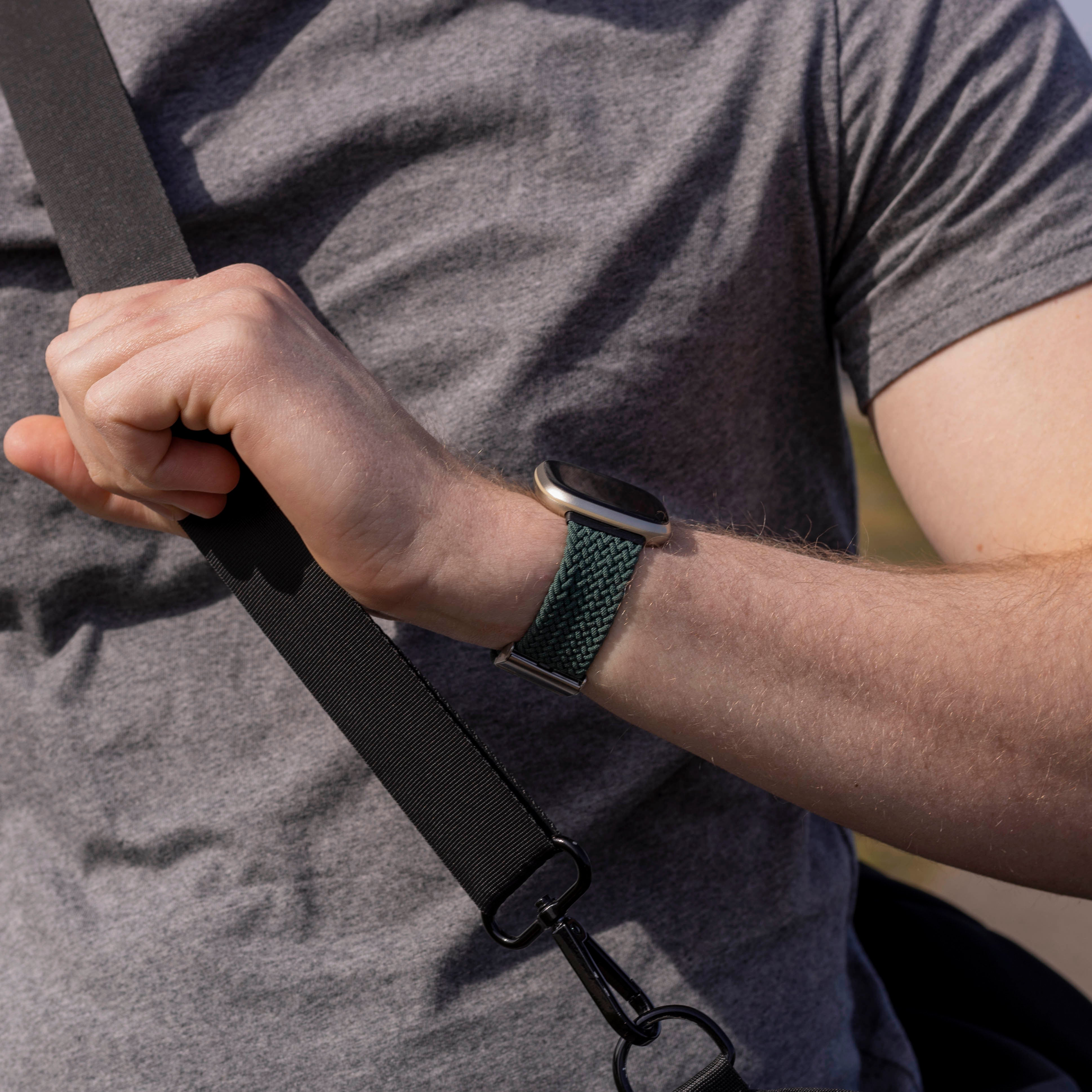 Der Arm eines Mannes mit grüner Uhr und grauem T-Shirt. Ein schwarzer Gurt wird von der Hand gehalten.