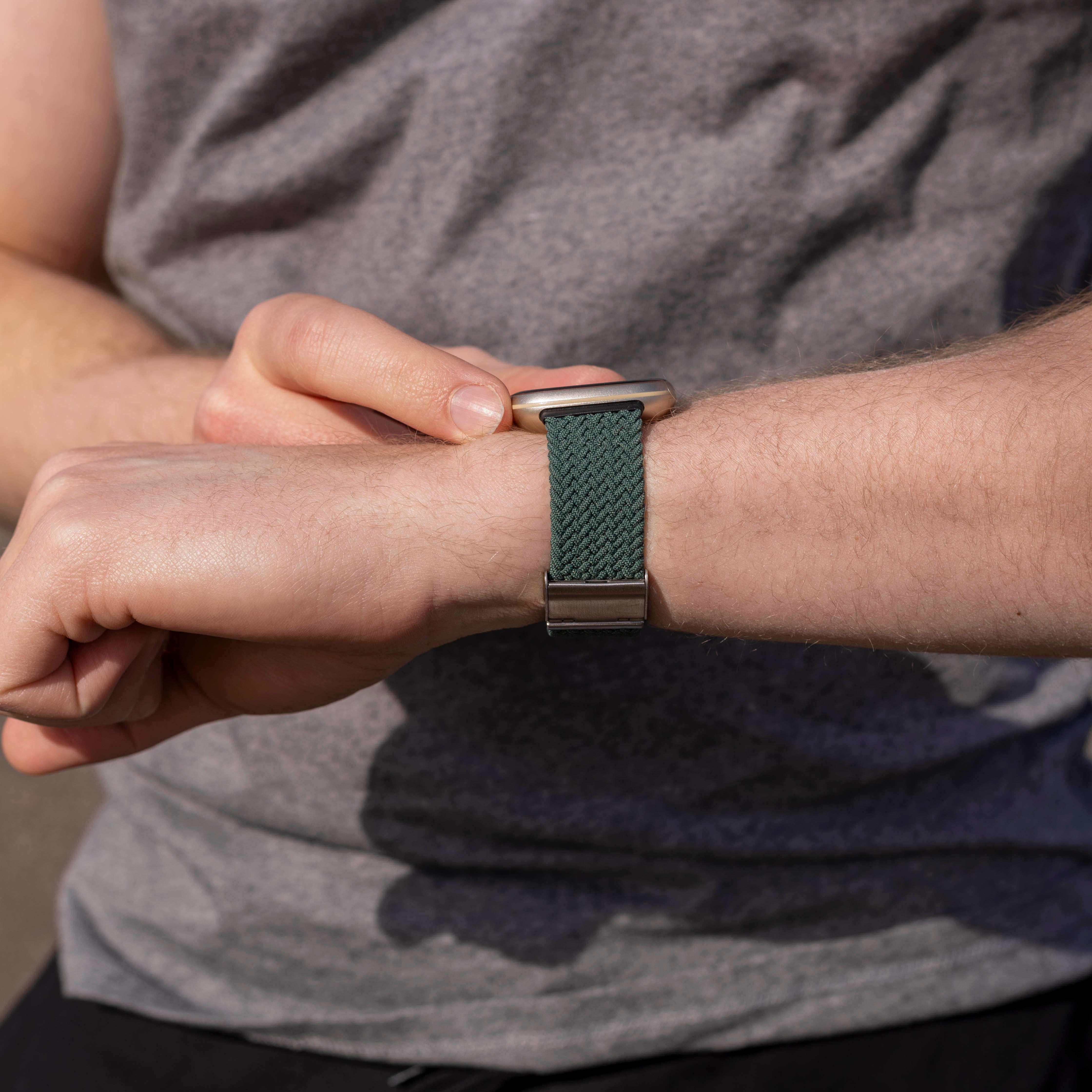 Arm mit Uhr. Die Uhr hat ein grünes geflochtenes Armband. Die Person trägt ein graues T-Shirt.