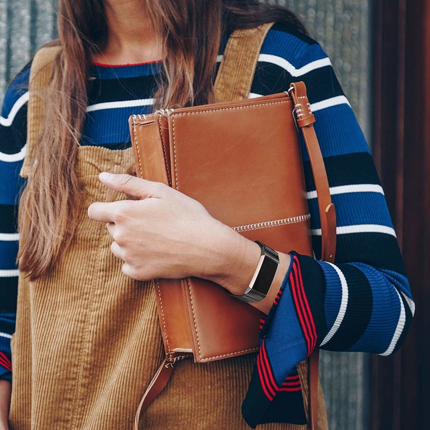 Frau hält eine braune Ledertasche, trägt einen blau gestreiften Pullover und Cord-Latzhose, mit einer schwarzen Smartwatch.