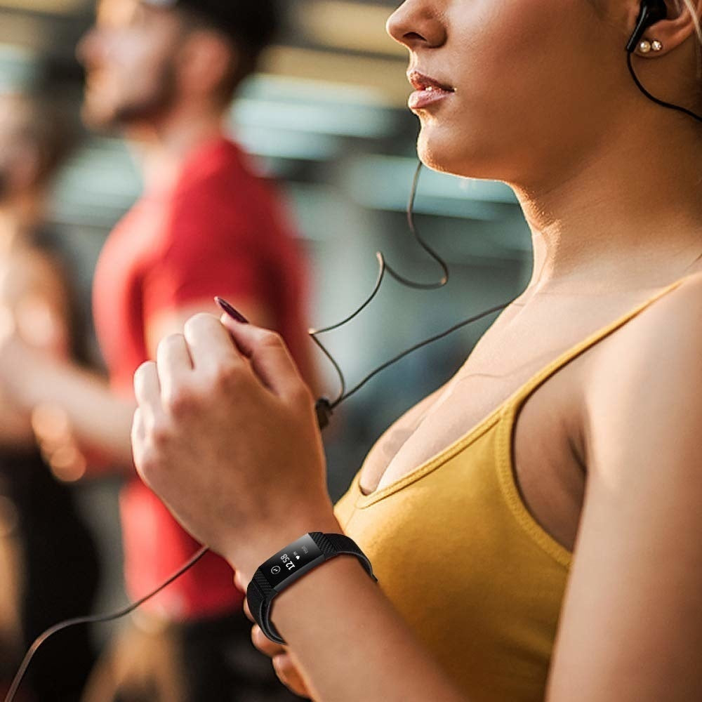 Eine Frau in einem gelben Oberteil mit Fitness-Tracker, die einen Kopfhörer hält. Im Hintergrund sind zwei verschwommene Männer in einem Fitnessstudio zu sehen.