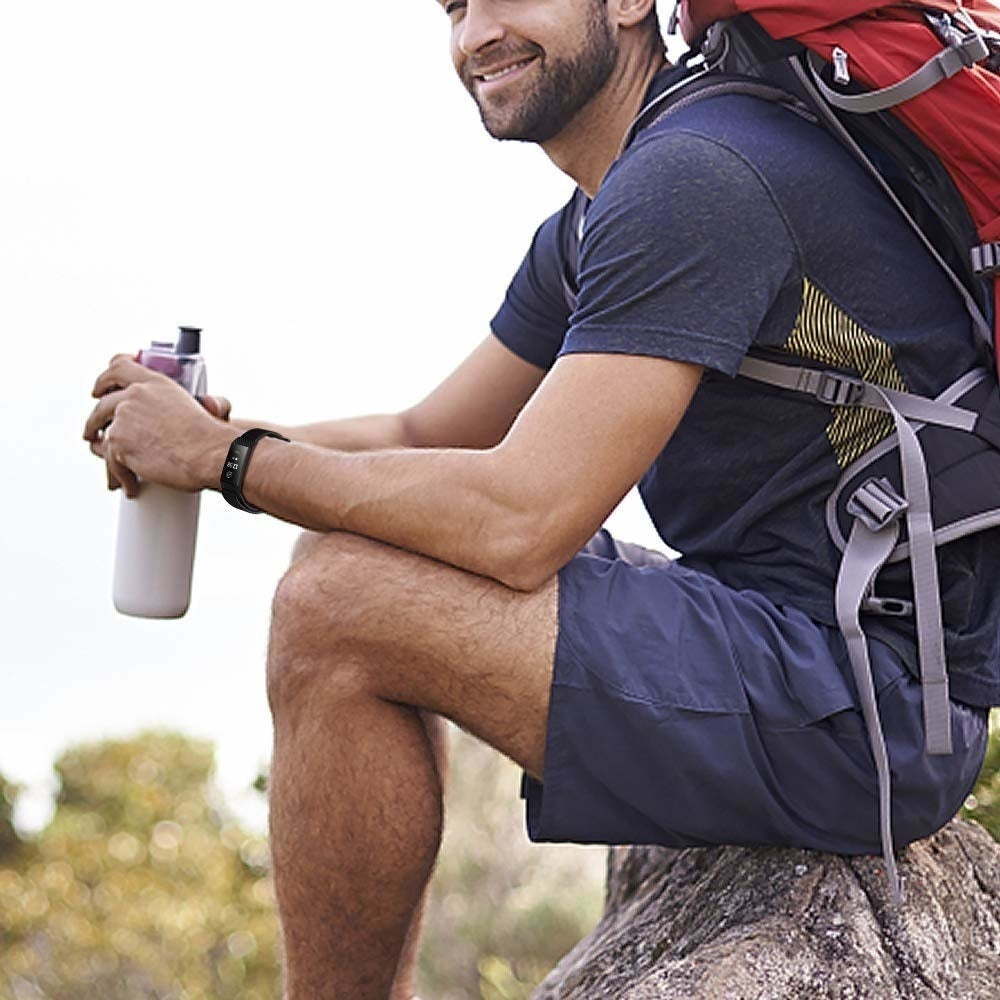 Ein Mann in Sportkleidung sitzt auf einem Felsen und hält eine Wasserflasche. Er trägt einen Rucksack und eine Uhr. Er lächelt in einer Umgebung im Freien.