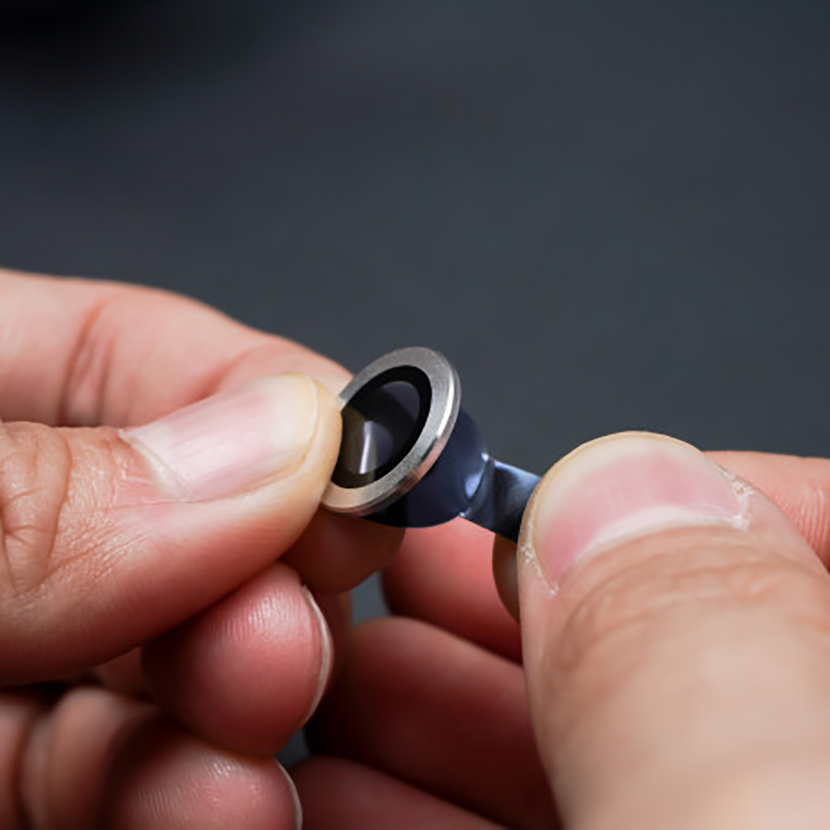 Handen die een kleine lens vasthouden met zilveren rand, zwart centrum en blauwe steel tegen een grijze achtergrond.