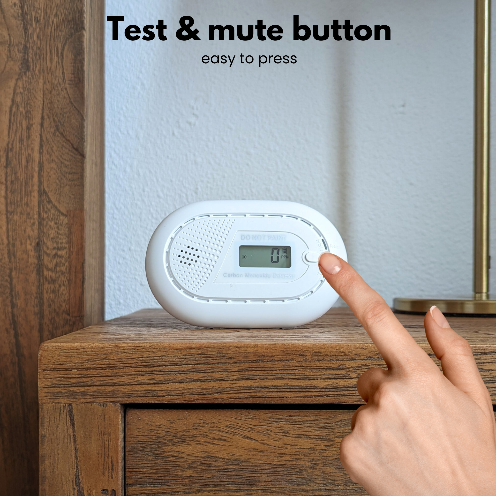 A hand presses the test button on a white carbon monoxide detector. The detector is on a wooden surface.