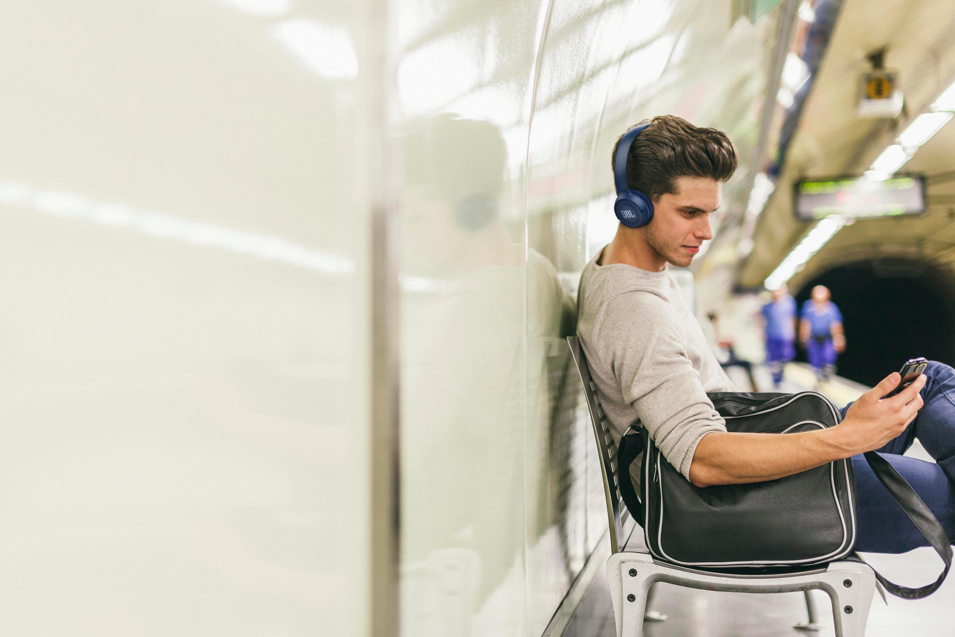 Man in blue headphones sits on a chair in the subway, looking at his phone. A bag is on the chair, and other people in the background.