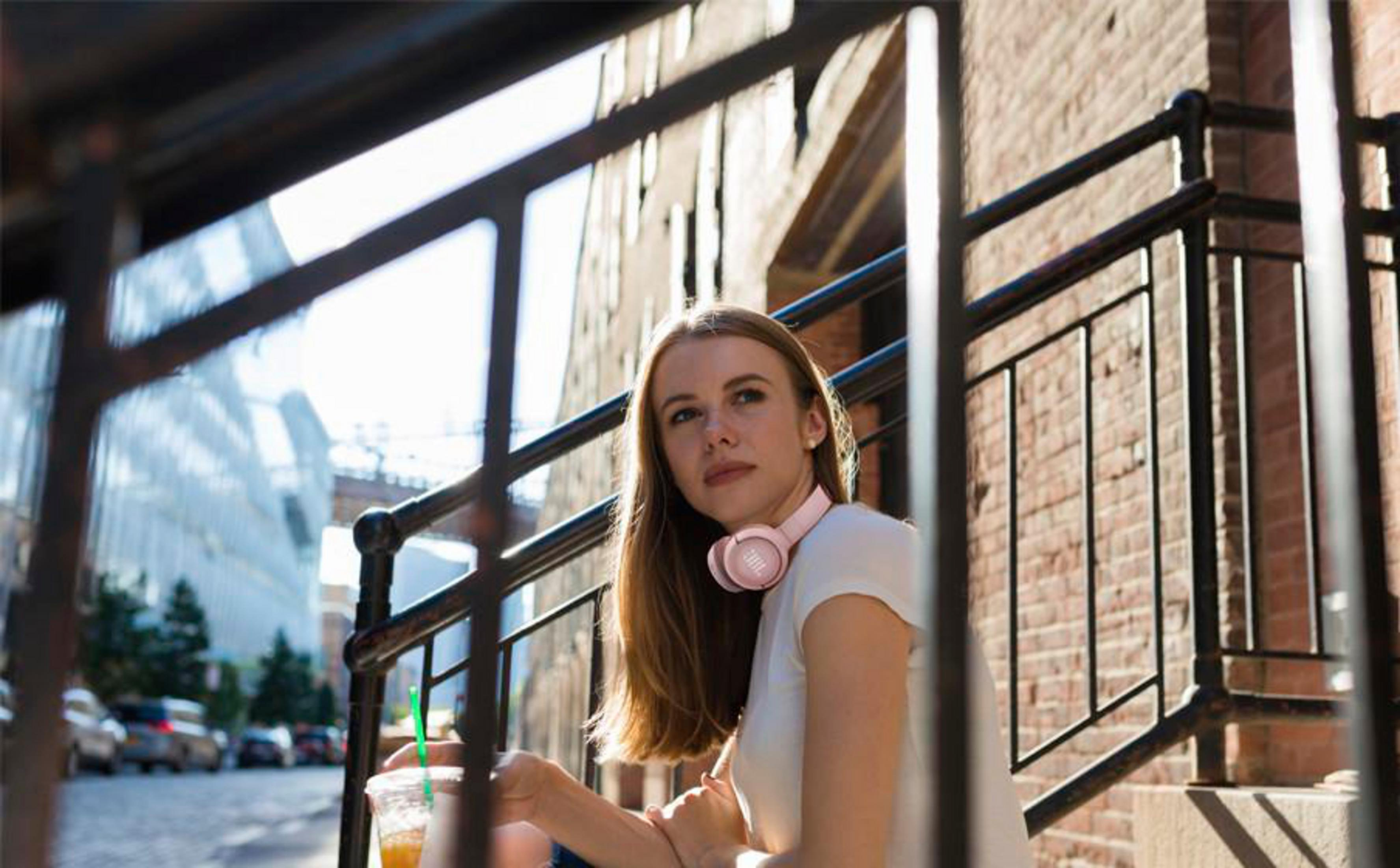 Woman with pink headphones sits on stairs, holding a drink. Brick wall background, black railing in the foreground.