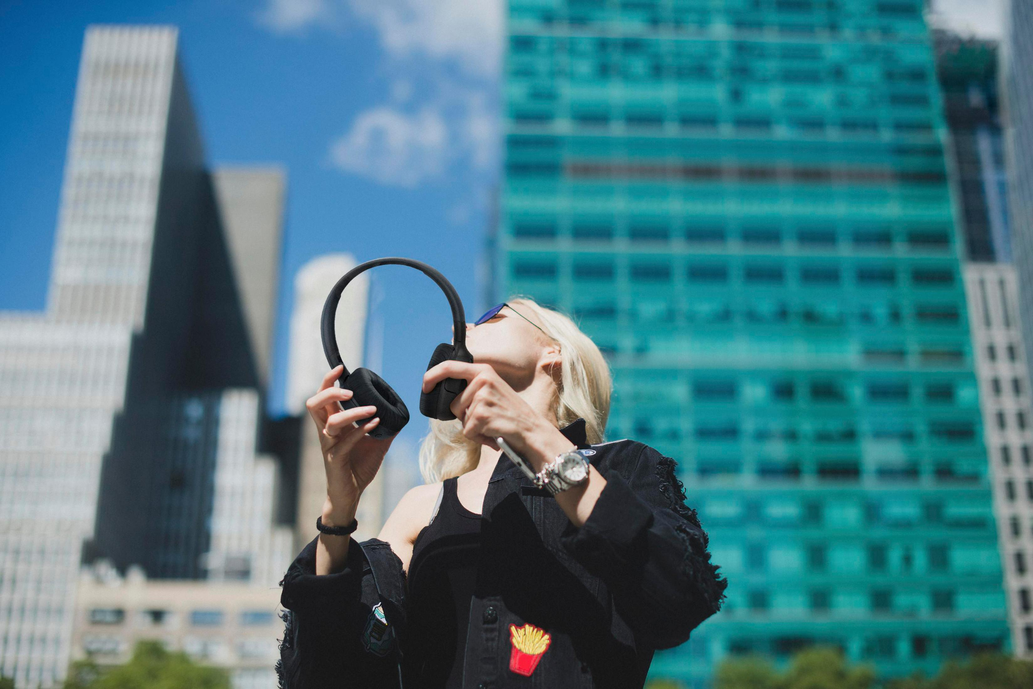 Blonde woman with headphones against city buildings. She holds the headphones up. Blue sky in background.