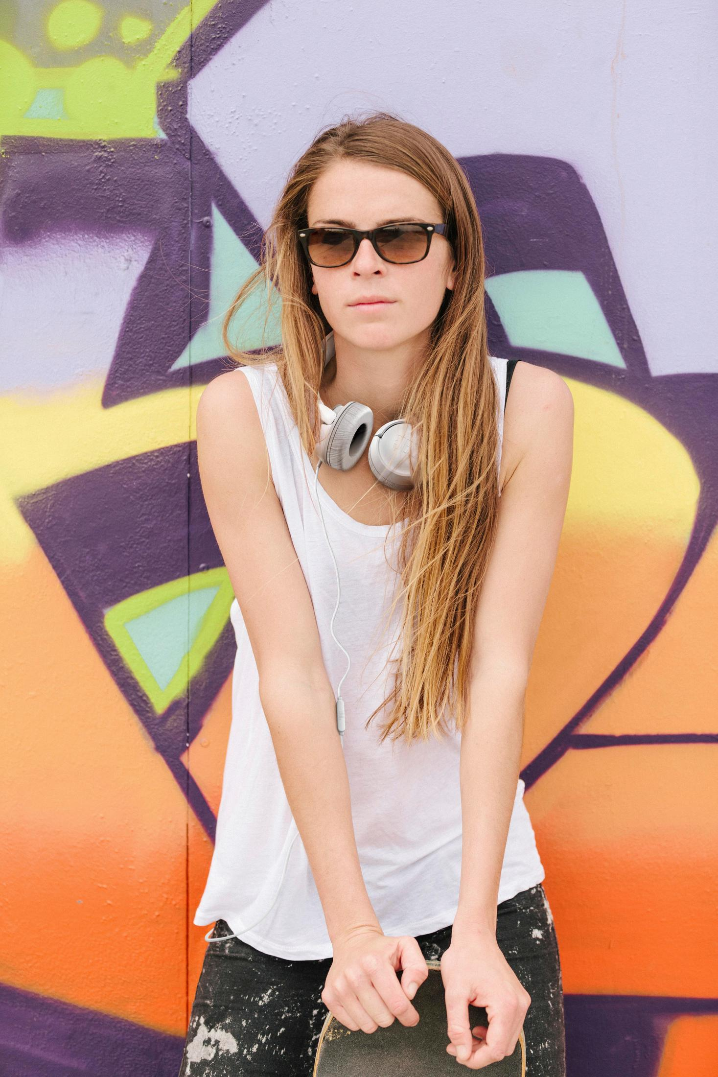 A young woman with sunglasses and headphones around her neck stands in front of a graffiti wall, holding a skateboard.