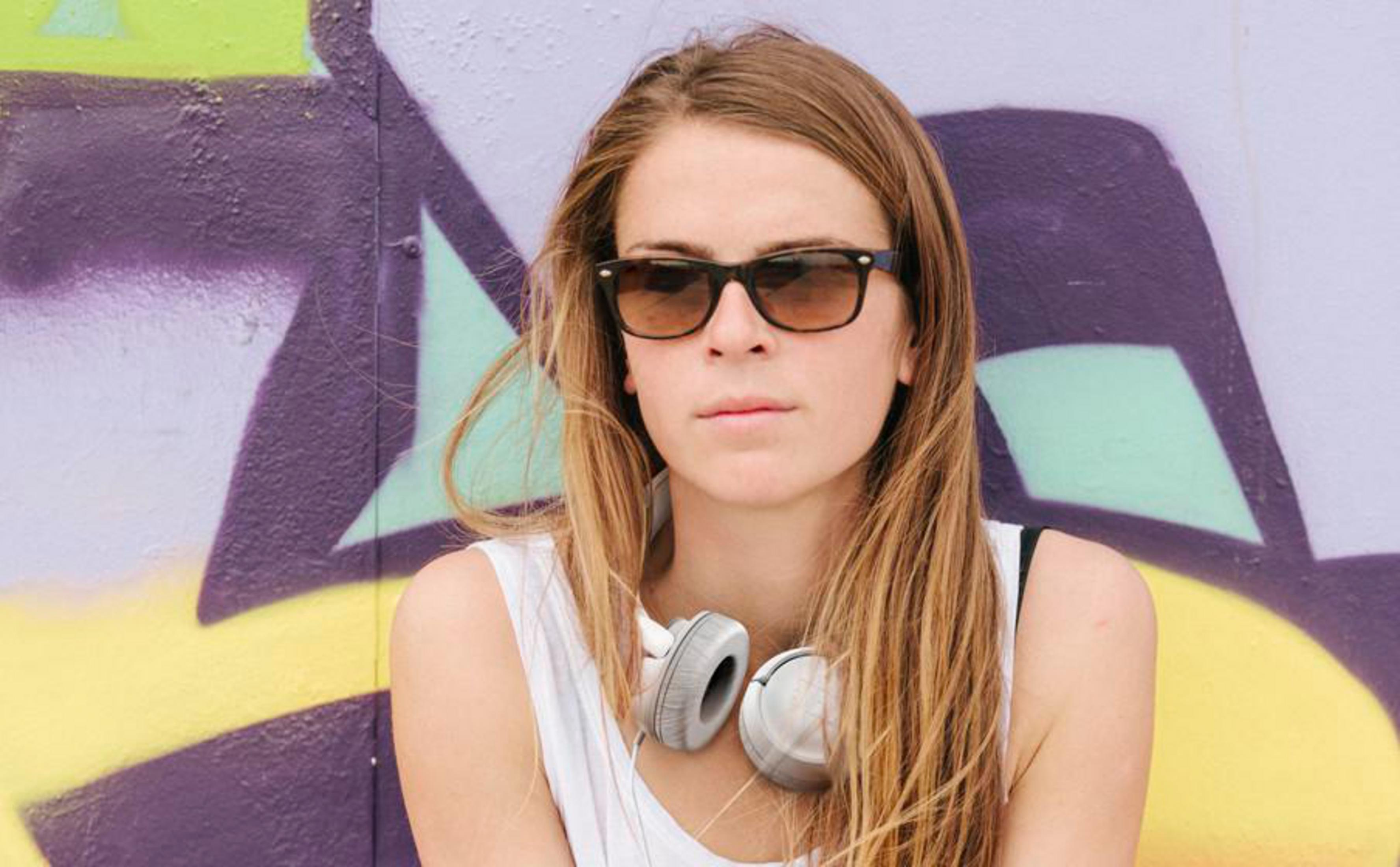 Young woman with sunglasses and headphones around her neck, stands in front of a colorful graffiti wall.
