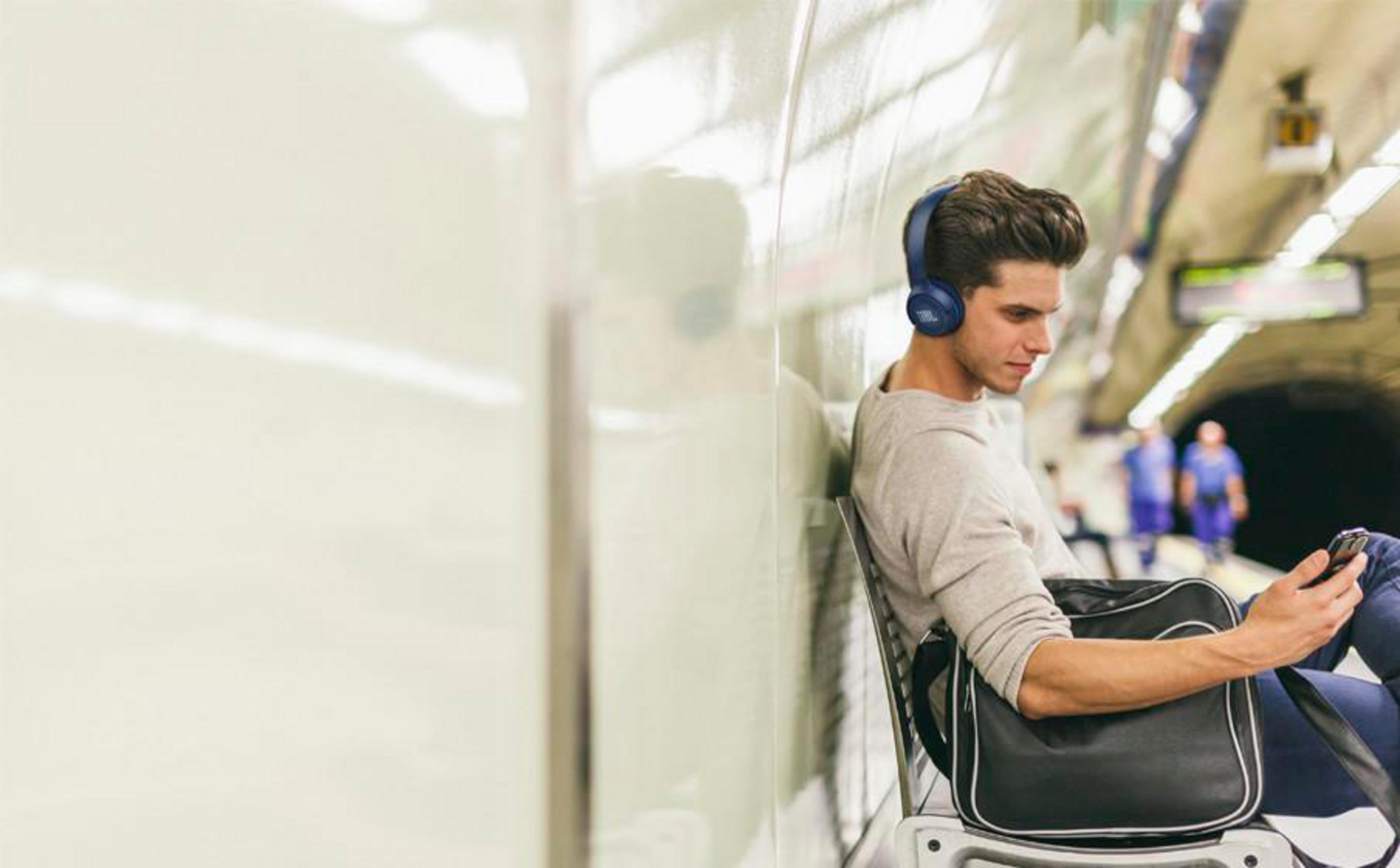 Man with headphones using a phone in a subway station. He is wearing a gray sweater and carrying a black bag.