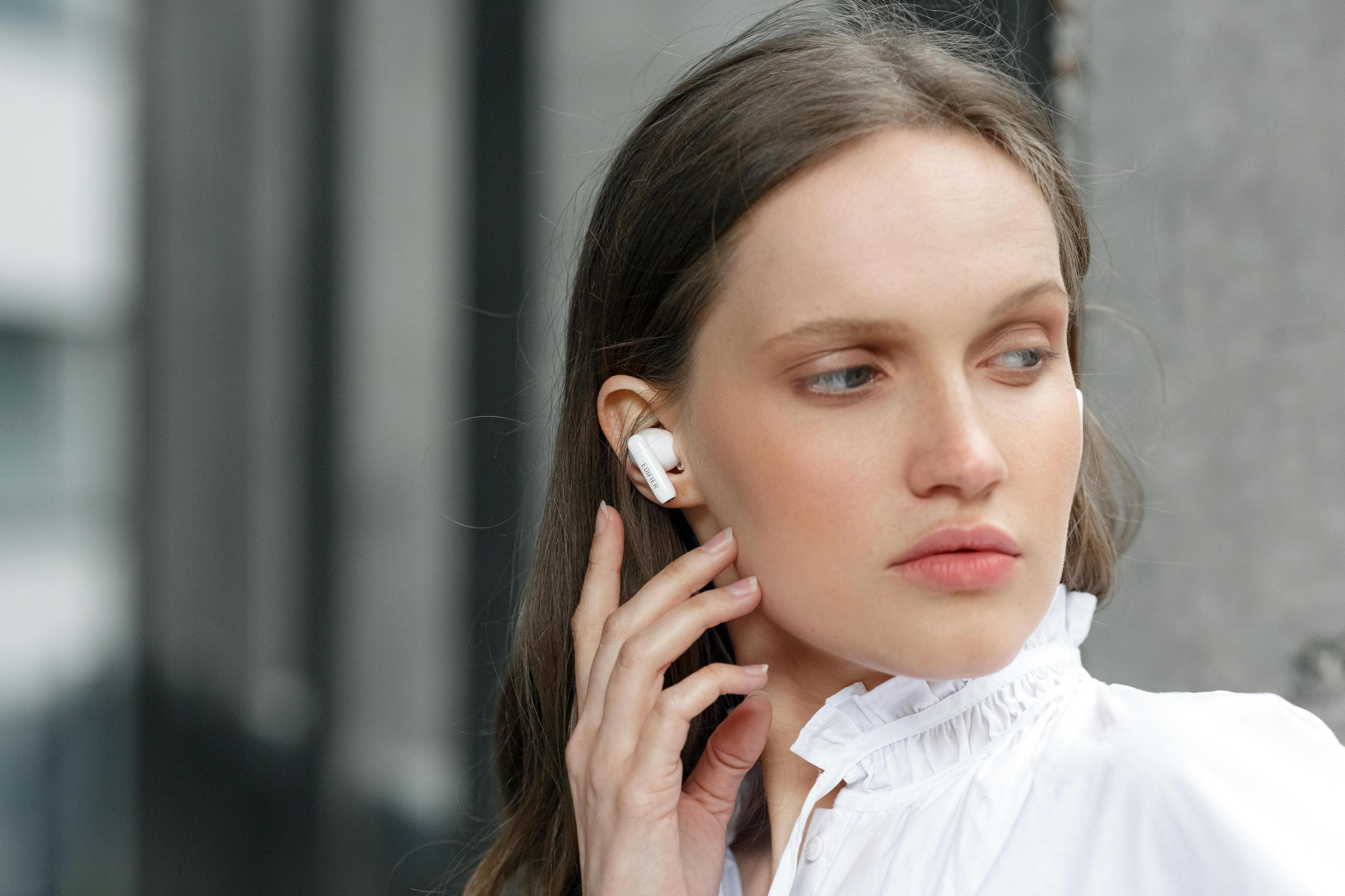 Kobieta w białych słuchawkach i białej koszuli dotyka twarzy. Widoczne jest szare tło.