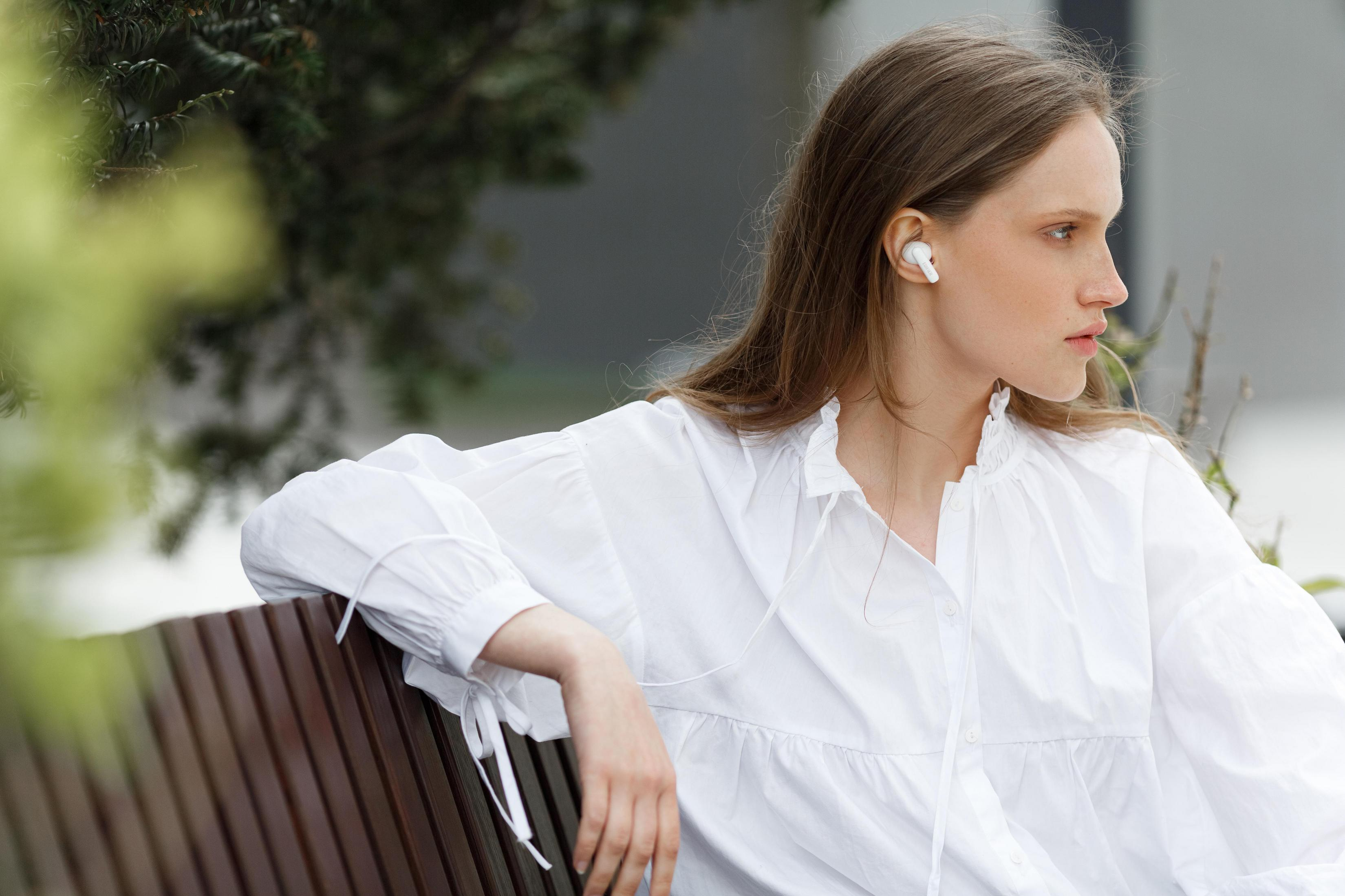 Kobieta w białej koszuli i słuchawkach siedzi na ławce. Tło przedstawia zieleń i budynek.