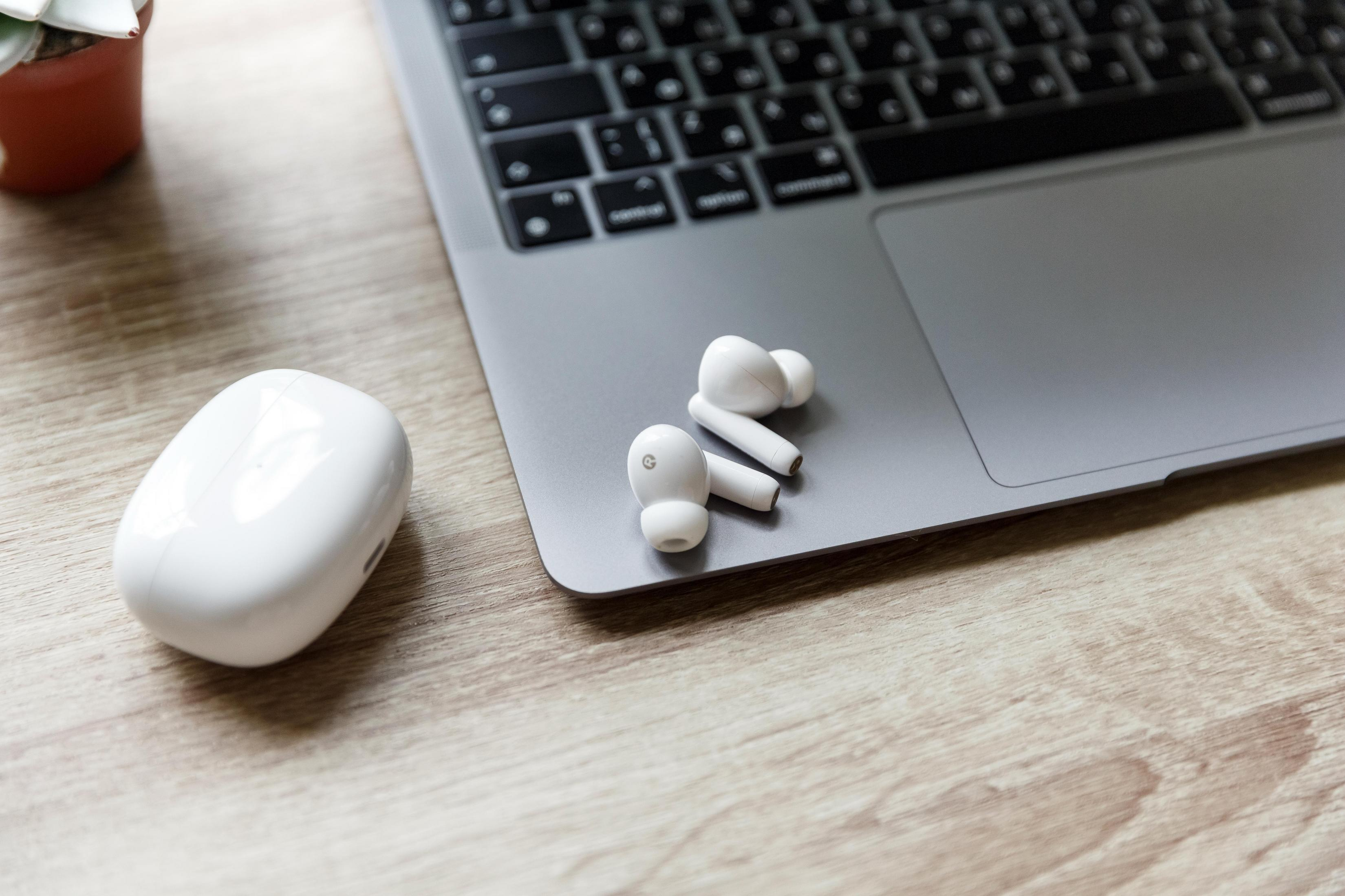 Laptop, białe słuchawki i etui ładujące na drewnianym biurku. Mała roślina w doniczce.