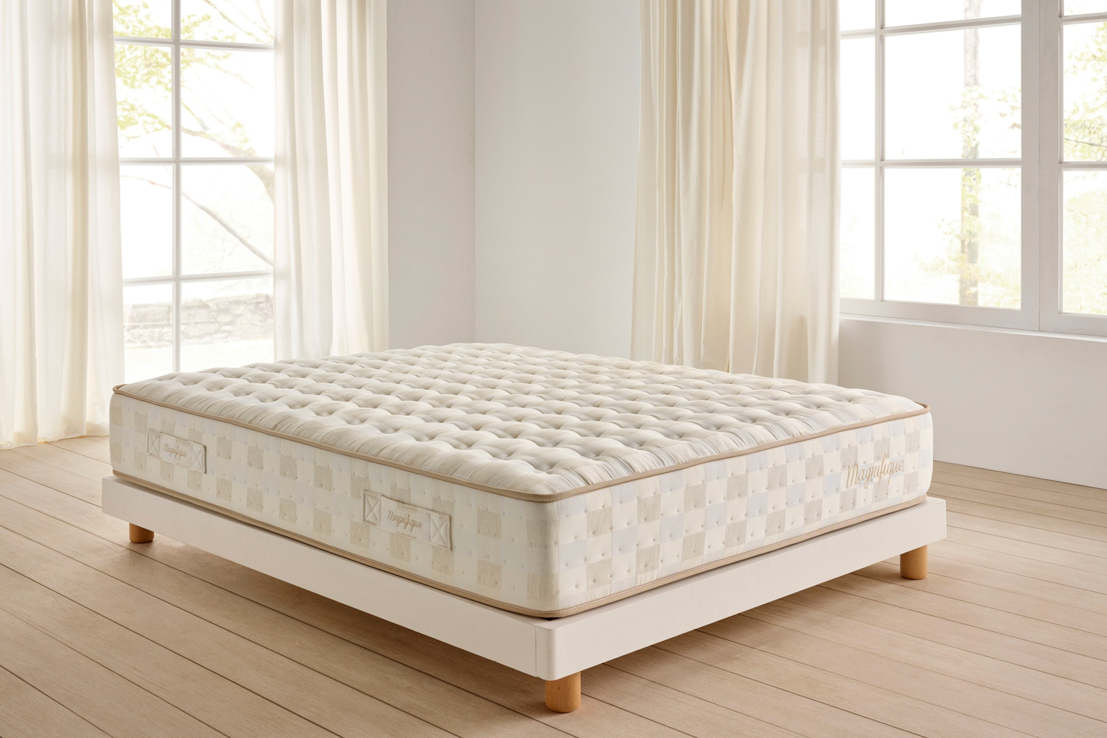 Un colchón grande de color crema sobre una cama con plataforma blanca. Las cortinas enmarcan una ventana brillante; suelo de madera.
