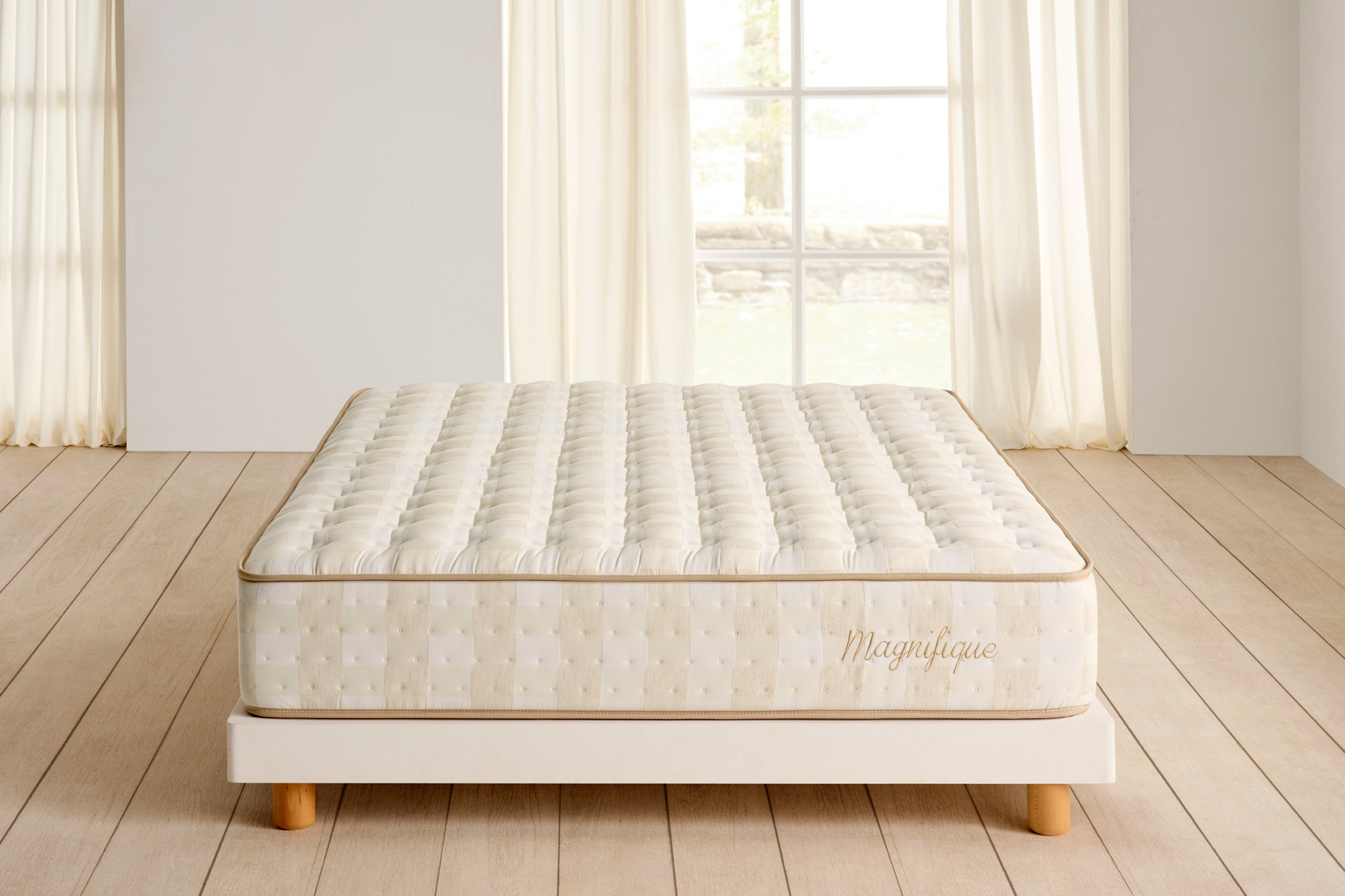 Un colchón crema sobre una estructura de cama blanca, en una habitación con ventana y cortinas. El colchón tiene la palabra Magnifique.