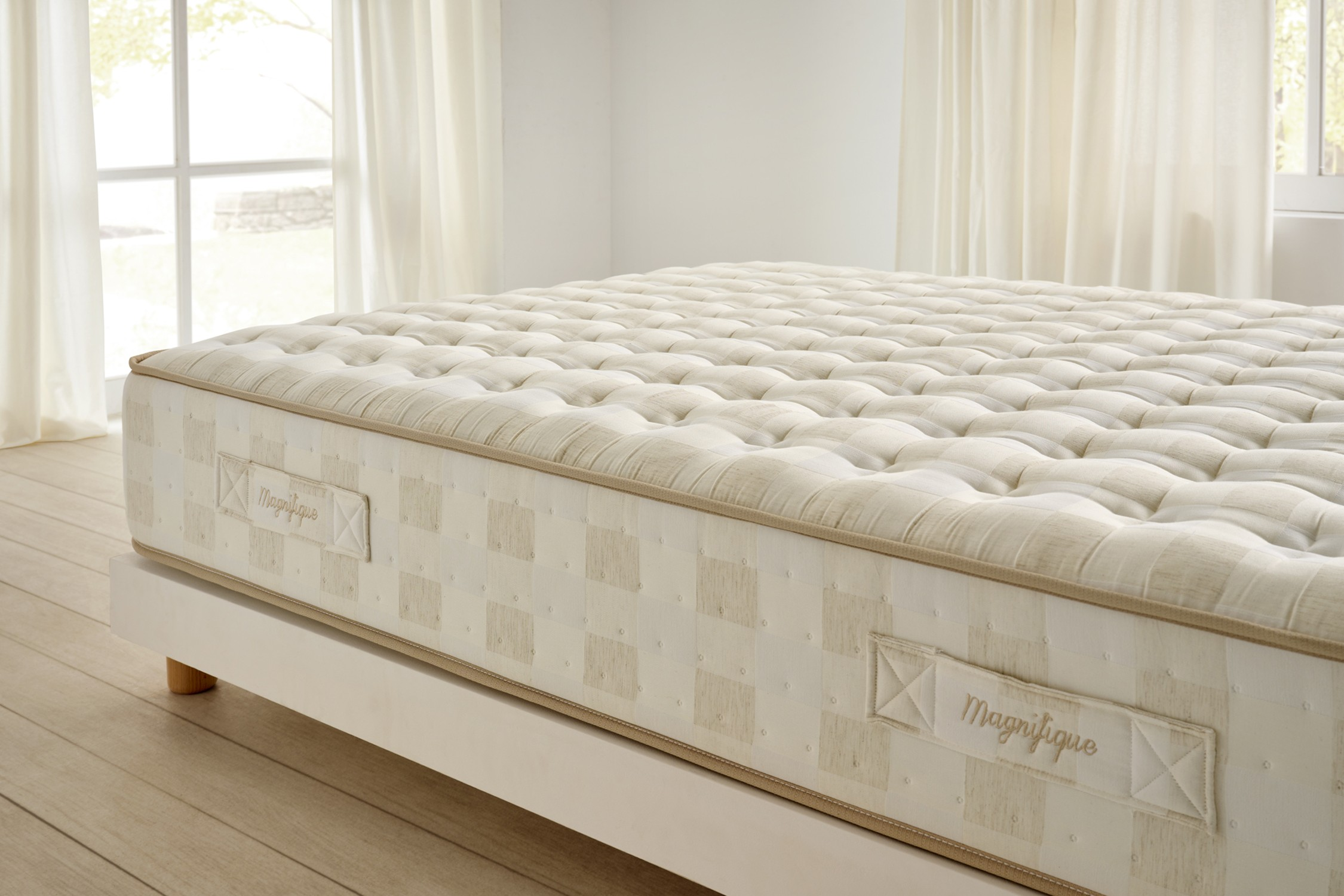 Un colchón afelpado y con mechones con la palabra 'Magnifique' en su lado. Se encuentra sobre una estructura de cama blanca en una habitación luminosa.