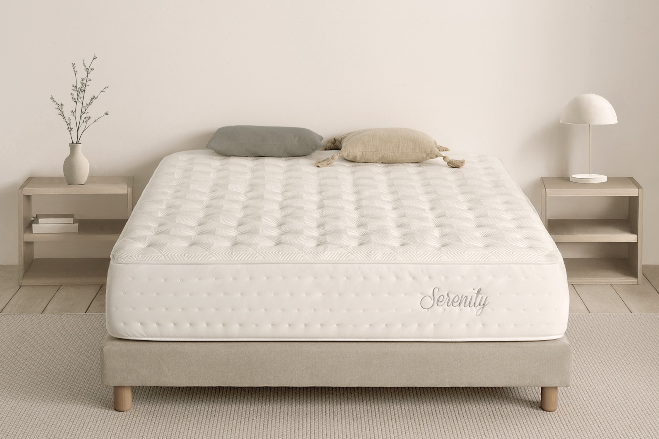 Escena de dormitorio con cama. Colchón blanco con almohadas, mesas auxiliares con lámpara y jarrón. Alfombra y pared beige.