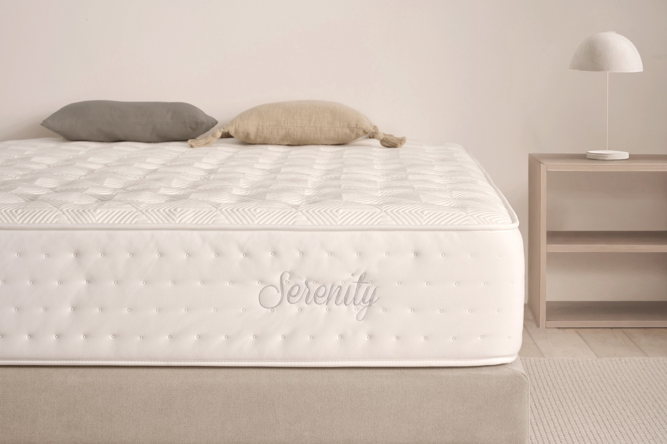 Un colchón blanco con 'Serenity' en el lateral, dos almohadas y una lámpara en una mesita de noche. Tonos neutros y una escena tranquila.